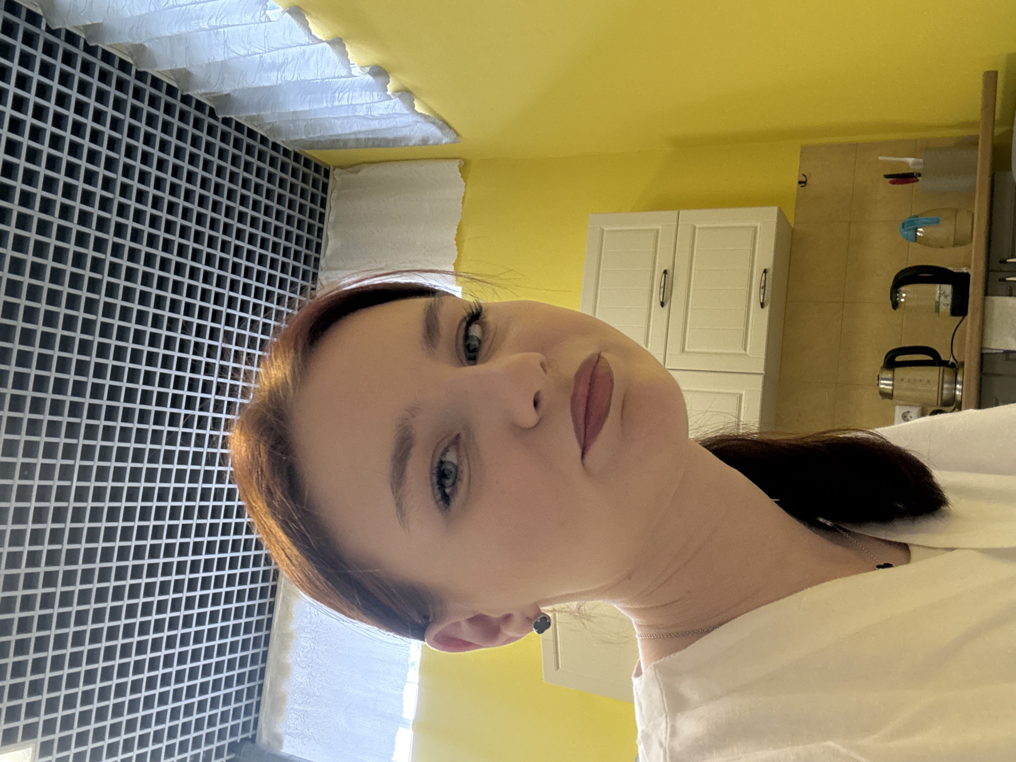 A medium shot of a woman with reddish-brown hair, fair skin, and blue eyes, possibly in an indoor environment. The woman has a serious expression on her face and is wearing a white garment. A black and white tiled wall is visible on the left, and a yellow wall and cabinets are in the background, giving the impression of a kitchen or similar interior space. Additional kitchen items can be seen on the right. Location: Unknown, Unknown.
