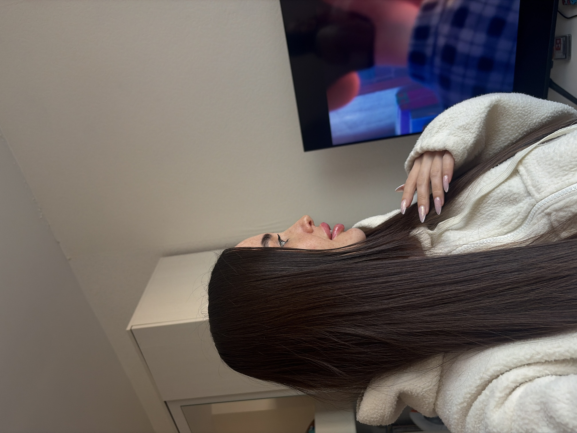 A woman with long brown hair and manicured nails is lying on a white, fluffy jacket in a room with white walls. A television is mounted on the wall in the background. The location is not specified.