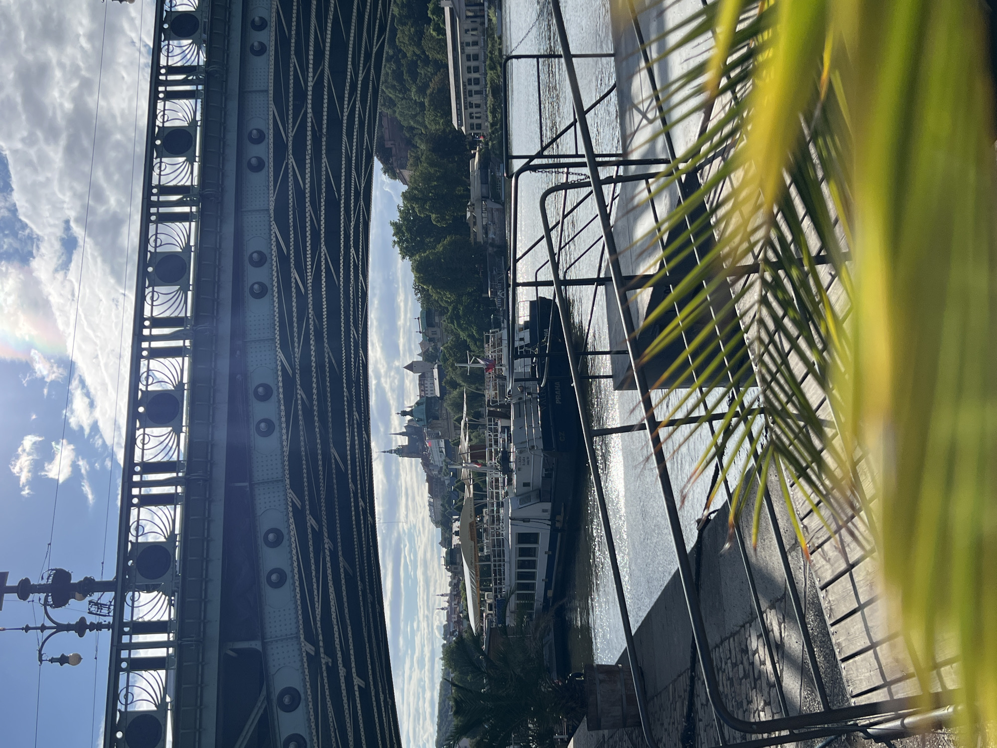The image captures a view from under a bridge, looking out towards a city skyline and a river. The bridge itself is a prominent feature, with its green metal structure and ornate railings visible in the foreground. Sunlight streams through gaps in the bridge structure, creating dappled patterns of light and shadow on the water and walkway below.
On the river, two white boats are moored. The boat on the left has a covered seating area on its upper deck. The boat on the right displays "CZ PRAHA" on its side, indicating it is in Prague. The city skyline in the background features historical buildings, including prominent spires and domes, suggesting a European setting. Trees line the riverbank, and a building with a sign that reads "RESTAURANT" is visible. In the sky, there are scattered clouds, and a faint rainbow can be seen to the left, near a street lamp attached to the bridge. In the immediate foreground, out-of-focus green foliage partially obscures the view, adding a layer of depth to the scene. The overall impression is one of a bright, sunny day in an urban waterfront setting.