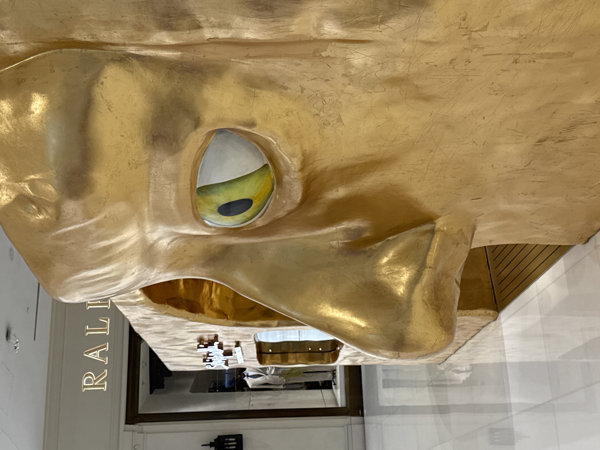 The image shows a close-up, side profile of a large, golden, abstract sculpture of a face. The sculpture features a prominent nose and an eye with a yellow and green iris and a black pupil, depicted in a stylized manner. The surface of the sculpture is textured with visible lines and scratches, reflecting light. In the background, a store with the sign "RALI" is visible, suggesting a retail environment, possibly a luxury department store. Through a glass display window, a mannequin dressed in a suit can be seen. The floor is tiled and highly reflective. This sculpture is located at The Dubai Mall in Dubai, United Arab Emirates.