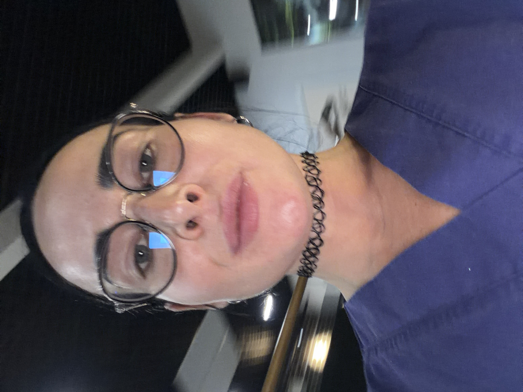 A medium close-up shot presents a person with fair skin, wearing round glasses and a black choker necklace. The person's face is prominently displayed, showing their nose, lips, and one visible ear adorned with a small earring. They are wearing a dark blue garment. The background consists of blurred lights and structural elements. The setting resembles an indoor location. The image does not definitively confirm that the location is Saint Petersburg, Russia, or provide specific details to determine the exact city location.