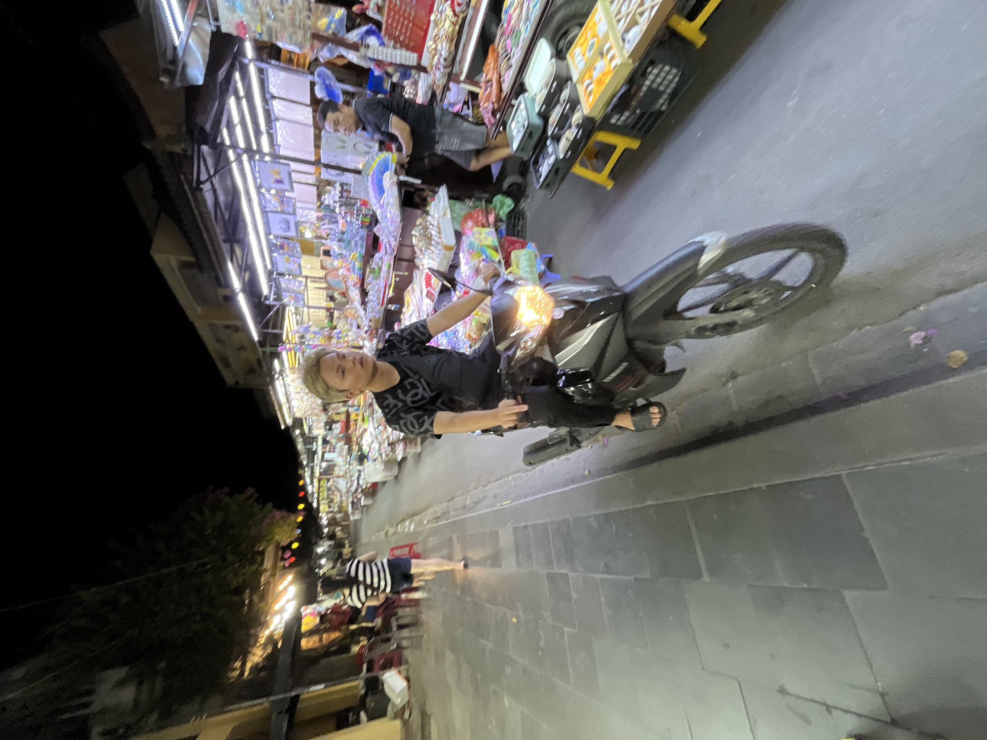 In Hội An, Vietnam, at night, a young man with blond hair is riding a black scooter on a paved street. He is wearing a black t-shirt with a patterned design and black pants, along with black flip-flops. The scooter has its headlights on, illuminating the road ahead. To the right of the scooter, a market stall is visible, displaying various goods. A man wearing a black t-shirt and gray shorts stands behind the stall, attending to customers or organizing merchandise. The stall is lit with bright fluorescent lights, and tables are laden with colorful items, including what appear to be souvenirs or crafts. In the background, more market stalls stretch along the street, with people browsing and vendors present. The street is lined with buildings, some with illuminated storefronts. Further down the street, another person, a woman wearing a black and white striped shirt and dark shorts, is walking away from the camera. The overall scene depicts a lively night market environment.