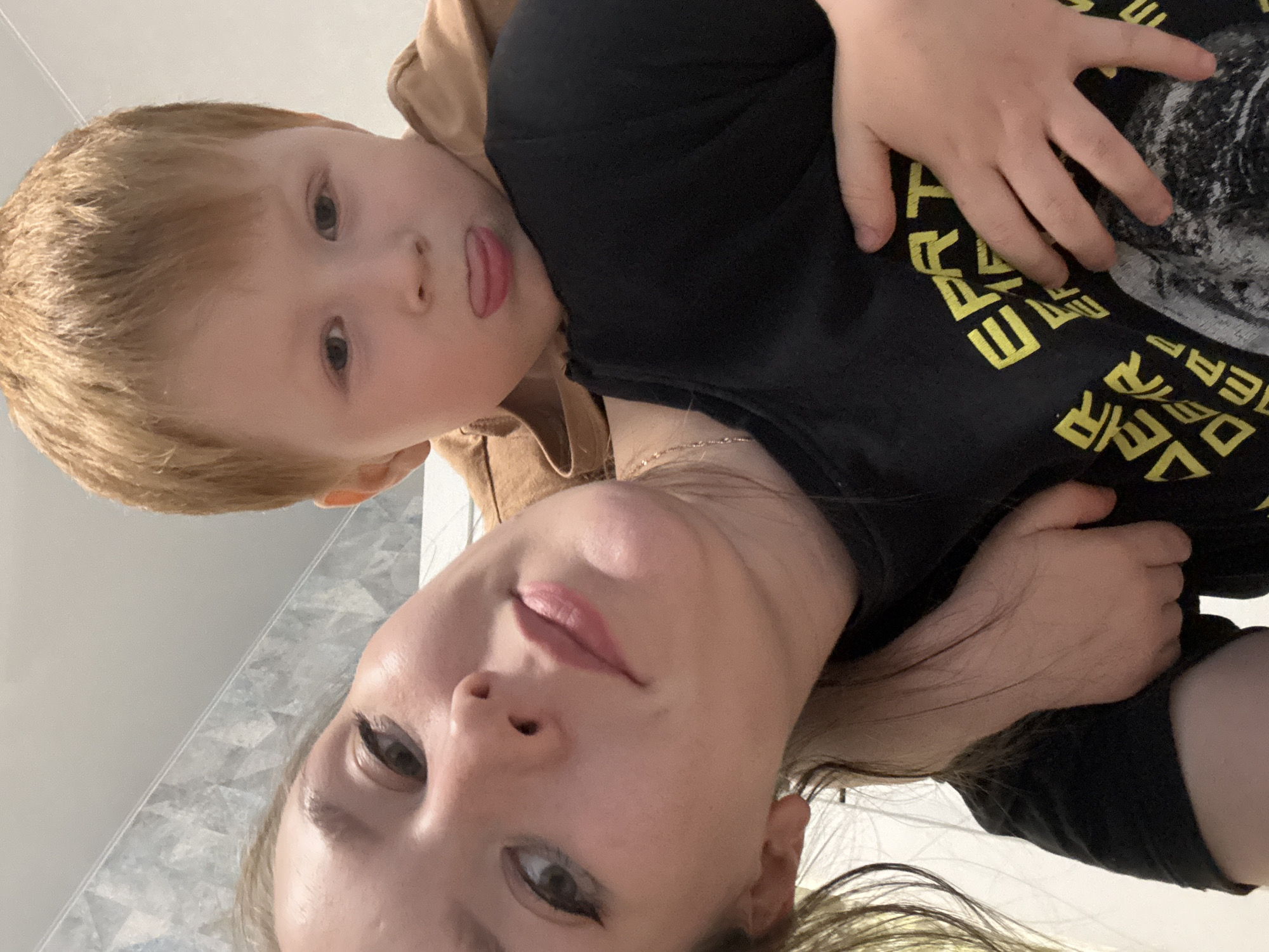 The image is oriented sideways, depicting a close-up, playful moment between a woman and a young child, likely a mother and son. The perspective suggests a selfie taken from a low angle.
The woman's face, positioned in the lower-right of the frame (when viewed upright), features light skin, dark eyes with subtle eyeliner, and light-colored lips. Her light brown hair flows around her. She wears a black t-shirt, and a thin gold chain is visible on her neck.
The child's face, in the upper-left (when viewed upright), has blonde hair, light skin, and dark eyes. His tongue is playfully sticking out. He is dressed in a light brown hoodie or jacket, and his hand rests on the woman's black t-shirt.
Yellow, stylized capital letters are printed on the black t-shirt. Visible fragments include "DEЯ", "EP", "Т", and "EЯ", suggesting Russian text. Given the Russian location context, these could be parts of a phrase like "ДЕРЗАЙ" (Dare).
The background is a light-colored, plain indoor surface, hinting at a home or public indoor setting. The scene conveys warmth and affection between the two individuals. The location context for this image is Izhevsk, Russia.