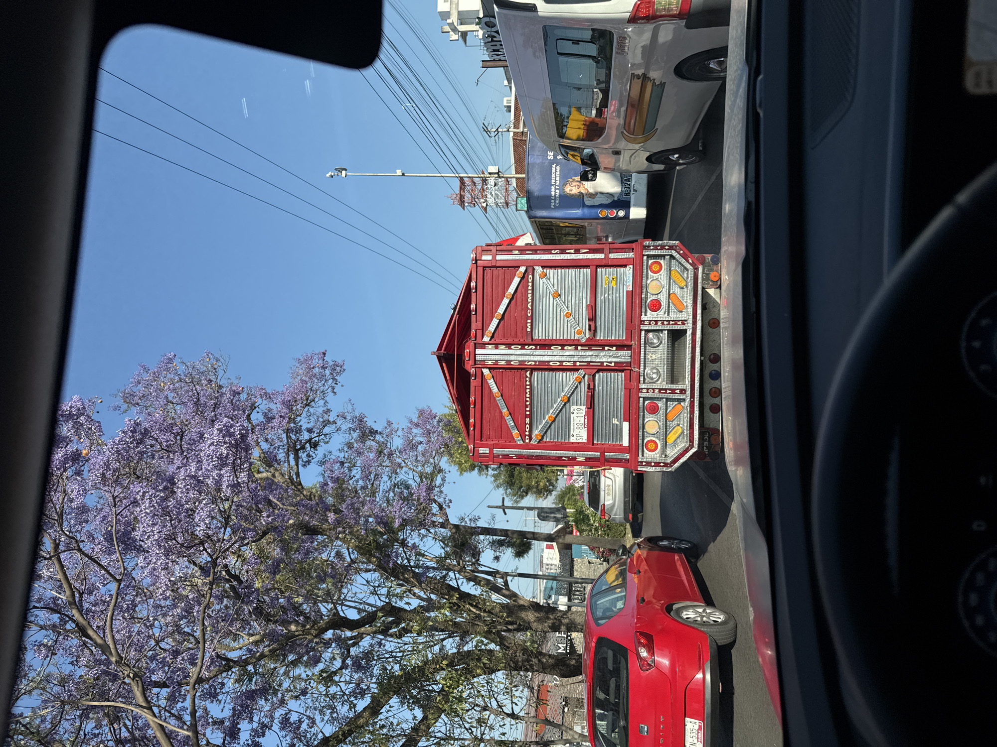 The image, taken from inside a vehicle, depicts a street scene in what is purportedly the city of Puebla, Mexico. A prominent feature is the back of a red truck, emblazoned with text that may contain details pertinent to its owner or operator. A red passenger car is to the right of the truck. There is a flowering tree to the left. Beyond the truck, other vehicles and a commercial van can be seen. Overhead power lines and a street light are present in the background. The sky is clear and blue. The presence of license plates and commercial advertising on the van suggests that this is a populated, urban environment.