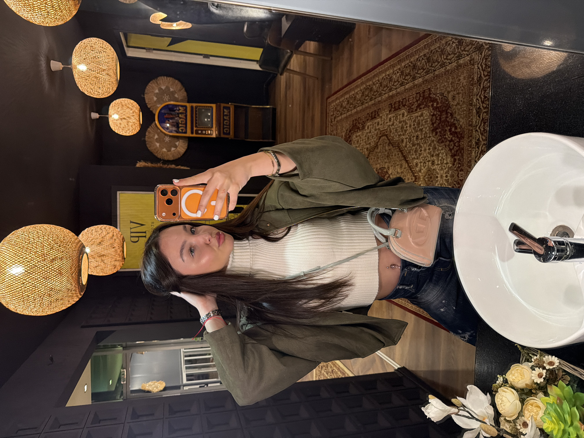 A young woman is taking a selfie in what appears to be a restroom or dressing room. She is wearing a white turtleneck crop top, dark wash jeans, and an olive green jacket. She has long dark hair and is holding an orange iPhone. A small pink purse hangs by her side. In the background, there are decorative light fixtures, a vintage-looking entertainment unit, and a patterned rug. A white sink with a chrome faucet and a bouquet of flowers are also visible. There is no indication that the scene is located in the city of Kulata, Bulgaria.