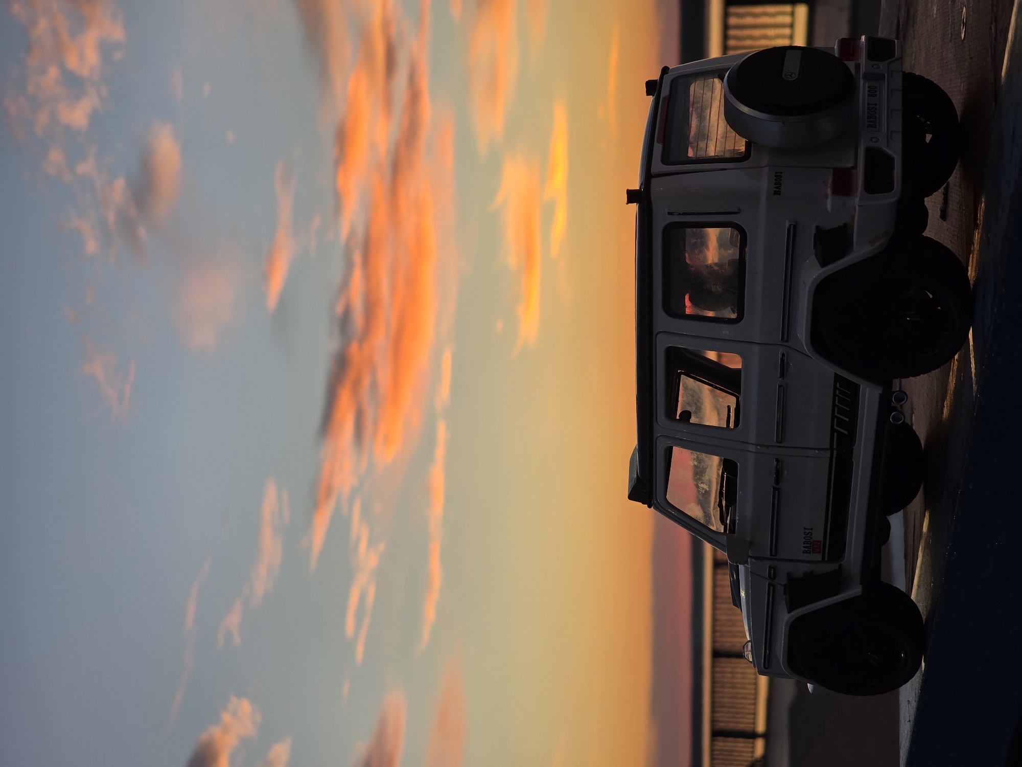 A toy replica of a white Mercedes-Benz G-Class is positioned on a dark blue surface, likely a railing or ledge, with a sunset sky in the background. The sky is filled with streaks of orange, pink, and blue clouds, indicating either sunrise or sunset. In the distance, a body of water, possibly a river or lake, is visible, with a bridge or embankment running across it. The toy vehicle is detailed, showing branding like "Brabus" and "V12". The lighting suggests it is late evening or early morning, casting long shadows. The environment appears to be outdoors, possibly near a waterfront in the city of Chaikovsky, Russia, given the prominent sunset and bridge-like structure. There are no people visible in the image.