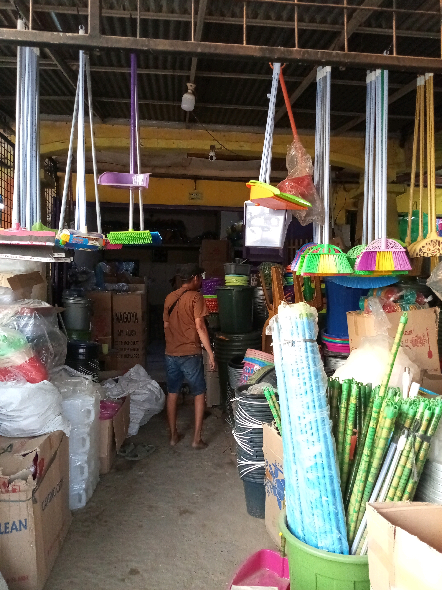 The image depicts a cluttered storage area or shop in Karangsatria, Indonesia, filled with cleaning supplies and other household items. Various brooms with colorful bristles are hanging from the ceiling. Several plastic buckets, some stacked, are visible on the floor. A man wearing a brown t-shirt and denim shorts is standing in the middle of the aisle, facing away from the camera. He appears to be looking at the items in the shop. In the foreground, there are stacks of plastic containers and cardboard boxes, some open and revealing their contents. The overall impression is of a busy, functional space stocked with a variety of goods.