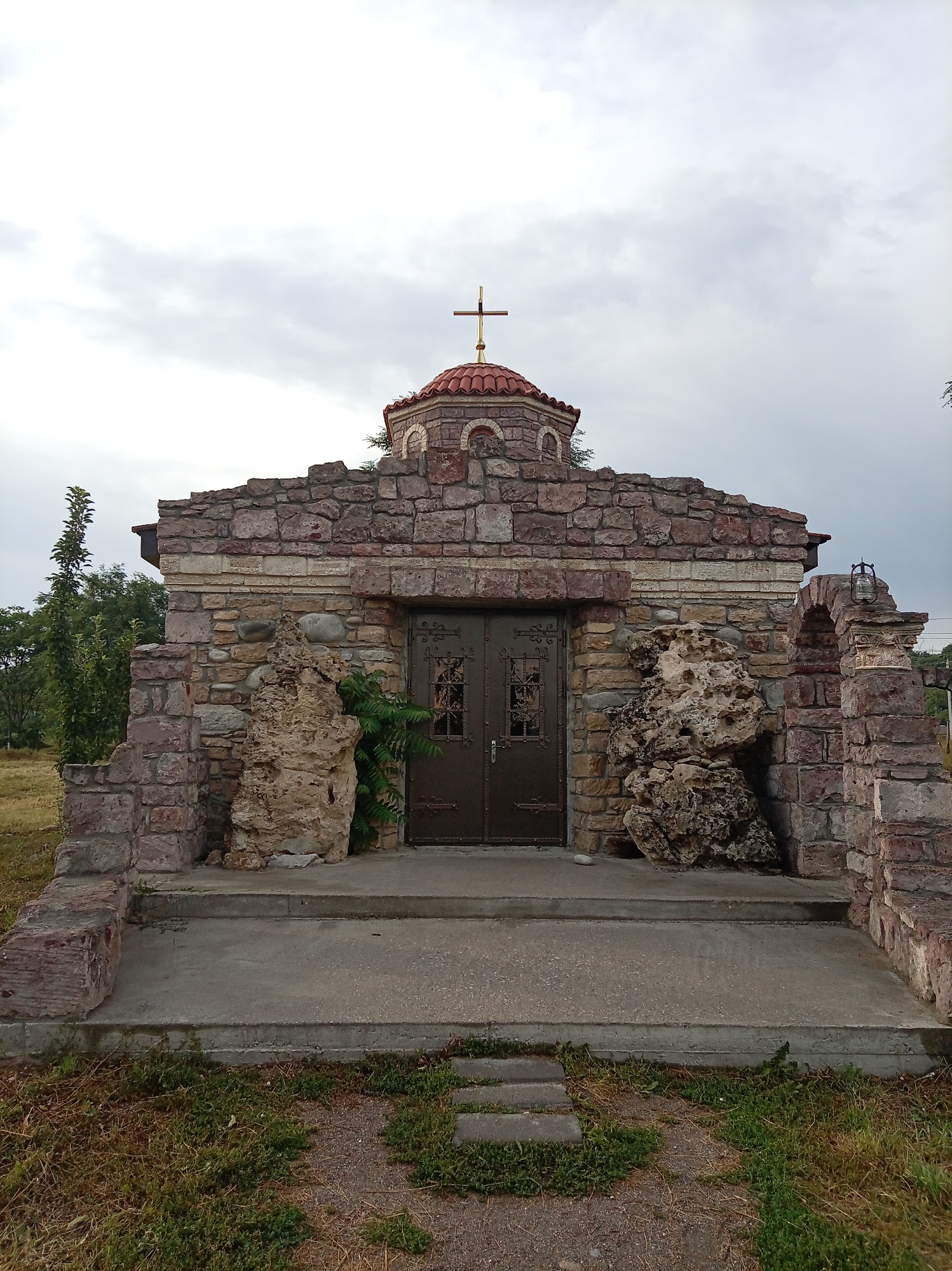 A small stone chapel with a red tiled dome topped by a gold cross stands under a cloudy sky. The chapel is constructed from rough-hewn stones of varying colors, primarily reddish-brown and grey. It has a double door made of dark metal with ornate carvings and small windows. Two large, irregular rock formations are placed on either side of the entrance, flanking the doors. To the left and right of the chapel, stone walls are visible, with foliage and trees in the background. A set of concrete steps leads up to the chapel entrance, and a path with stepping stones is present in the foreground. The scene appears to be outdoors, in a rural or semi-rural setting. The location is identified as being in the city of Yessentuki, Russia.