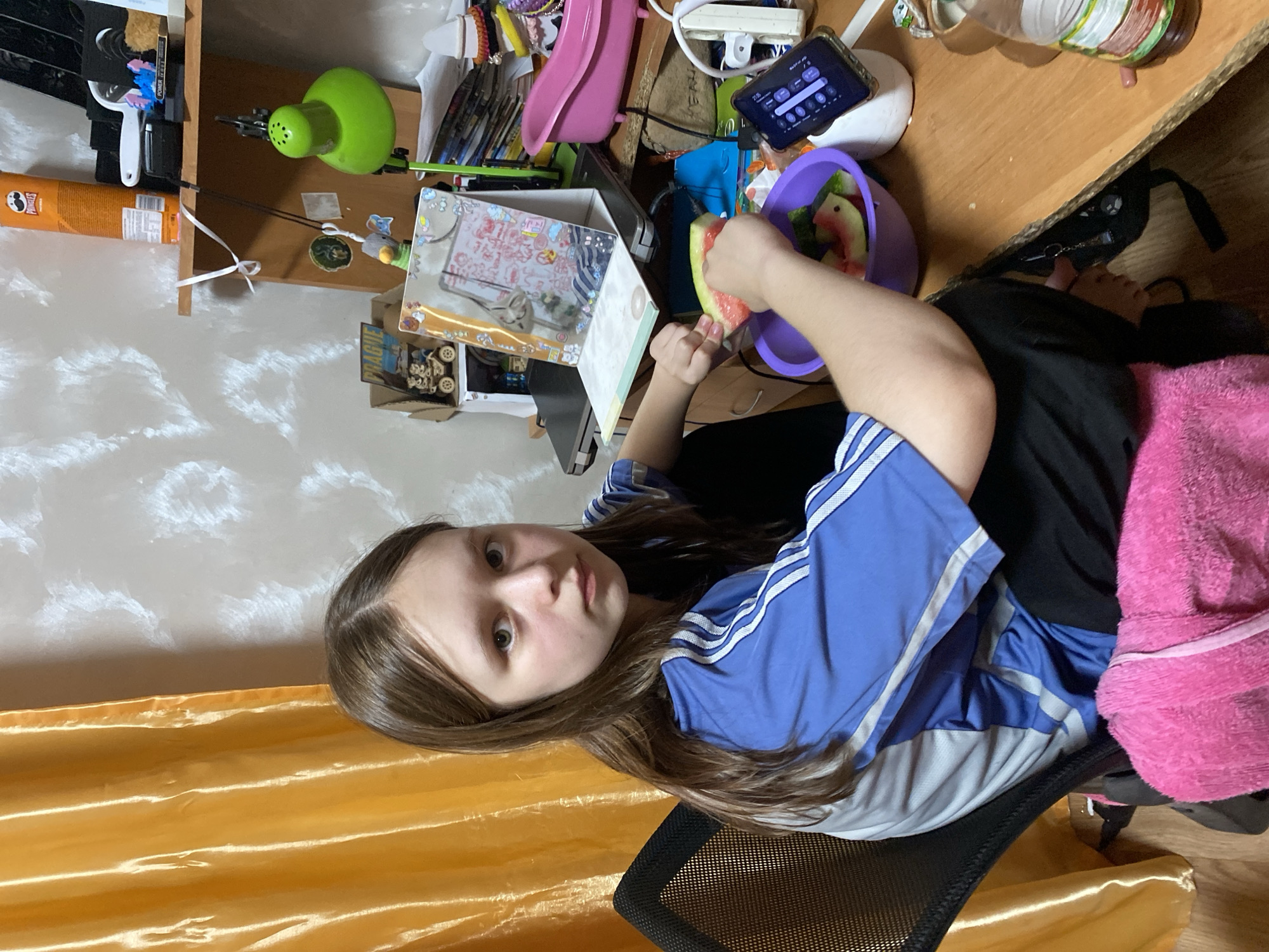A young girl with long brown hair is sitting at a desk, holding a slice of watermelon. She is wearing a blue and white striped t-shirt and black pants. She appears to be looking at the camera with a neutral expression. The desk is cluttered with various items, including a green lamp, a cell phone, a purple bowl with watermelon slices, and a box with stickers. A pink blanket is draped over the back of the chair she is sitting in. The background shows a wall with a patterned wallpaper and a yellow curtain. The lighting suggests it is daytime. The location is Chornomorsk, Ukraine.