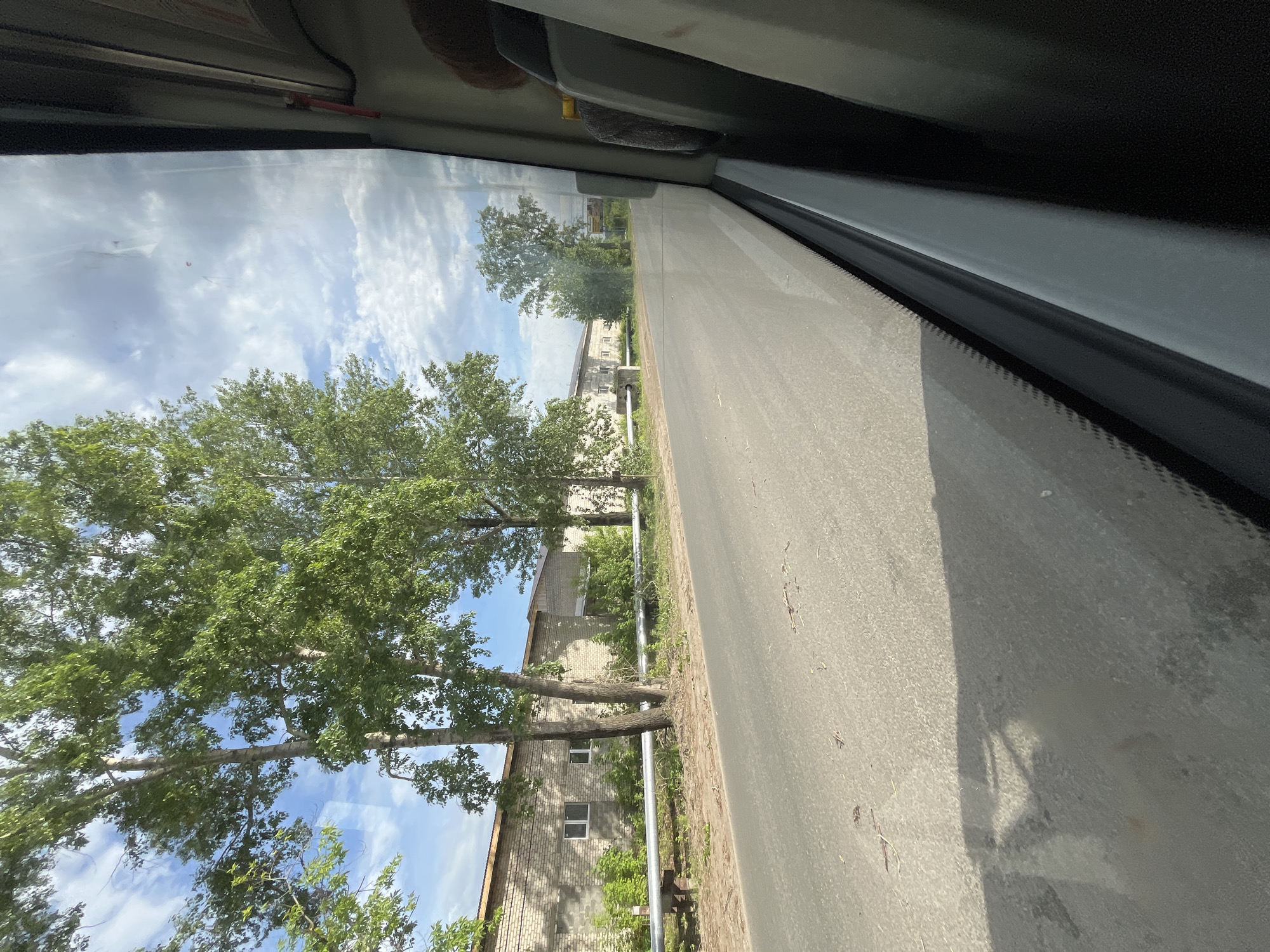 The image depicts a view from inside a vehicle, likely a bus or car, looking out the side window. A road is visible outside, running alongside a row of trees and buildings. The buildings appear to be low-rise, possibly residential or commercial structures. The sky is partly cloudy. The location is likely in Chaikovsky, Russia, given the request. The image is taken from a slightly elevated perspective, and the focus is on the outside scenery.