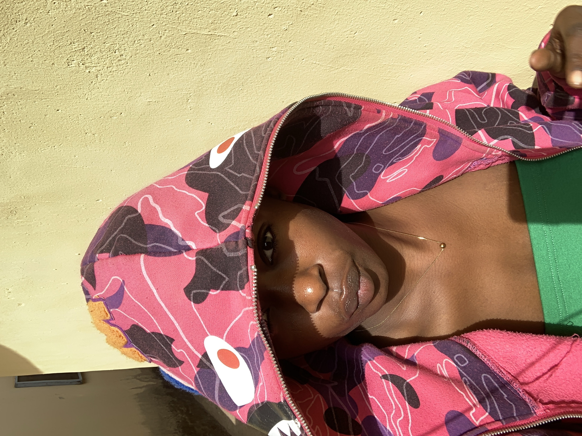 A medium shot depicts a Black woman, wearing a green top and a zip-up hooded sweatshirt with a pink and purple camouflage pattern and a graphic of an animal's face, positioned against a yellow wall. The woman's face is centrally framed with the hood pulled partially over her head, casting shadows across her face. She is looking directly at the camera. She is wearing a delicate gold chain necklace. The background is a plain yellow wall. The location in the city of Rafin Par, Nigeria, cannot be determined from this image.