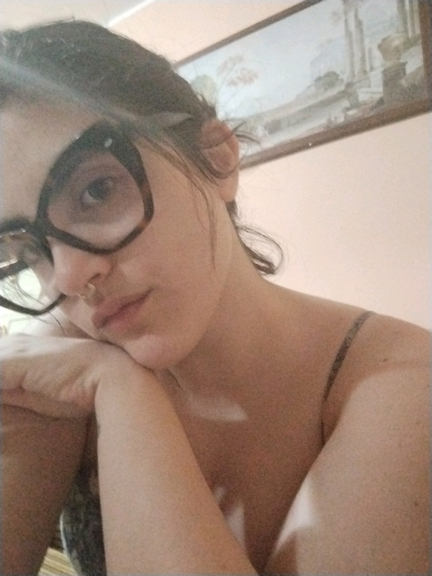A close-up, low-angle shot shows a young woman with dark hair pulled back, wearing large, dark-rimmed glasses and a septum piercing. She is resting her head on her hands, with her left arm bent and her right arm obscured. Her expression is neutral, and she appears to be looking slightly to her left. She is wearing a thin-strapped top, revealing her shoulder and upper chest. The background is a lightly textured pink wall with a framed picture hanging on it. The overall lighting is soft and warm, suggesting an indoor setting. The location is identified as Luzzi, Italy.