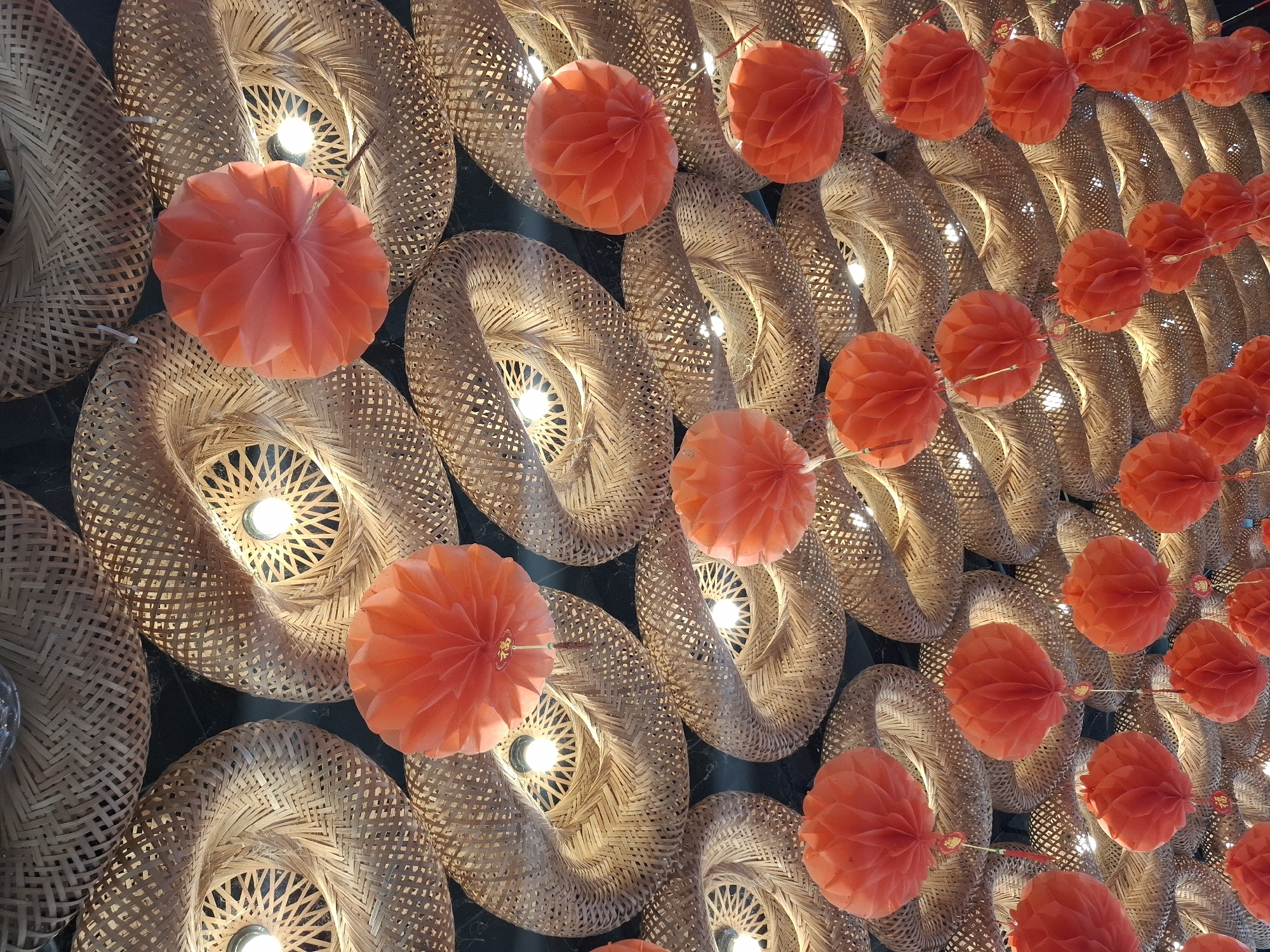 The image depicts an overhead view of a ceiling decorated with a complex arrangement of woven bamboo lampshades and hanging red paper lanterns. The bamboo shades are circular with intricate woven patterns, and each contains a lit bulb that casts a warm glow. The red paper lanterns are spherical and appear to be made of folded paper, creating a honeycomb-like texture. Some lanterns have small golden charms attached. The overall impression is one of festive and cultural decoration, likely in a restaurant or event space. This scene is located at a cafe on Trần Phú Street in Nha Trang, Vietnam.