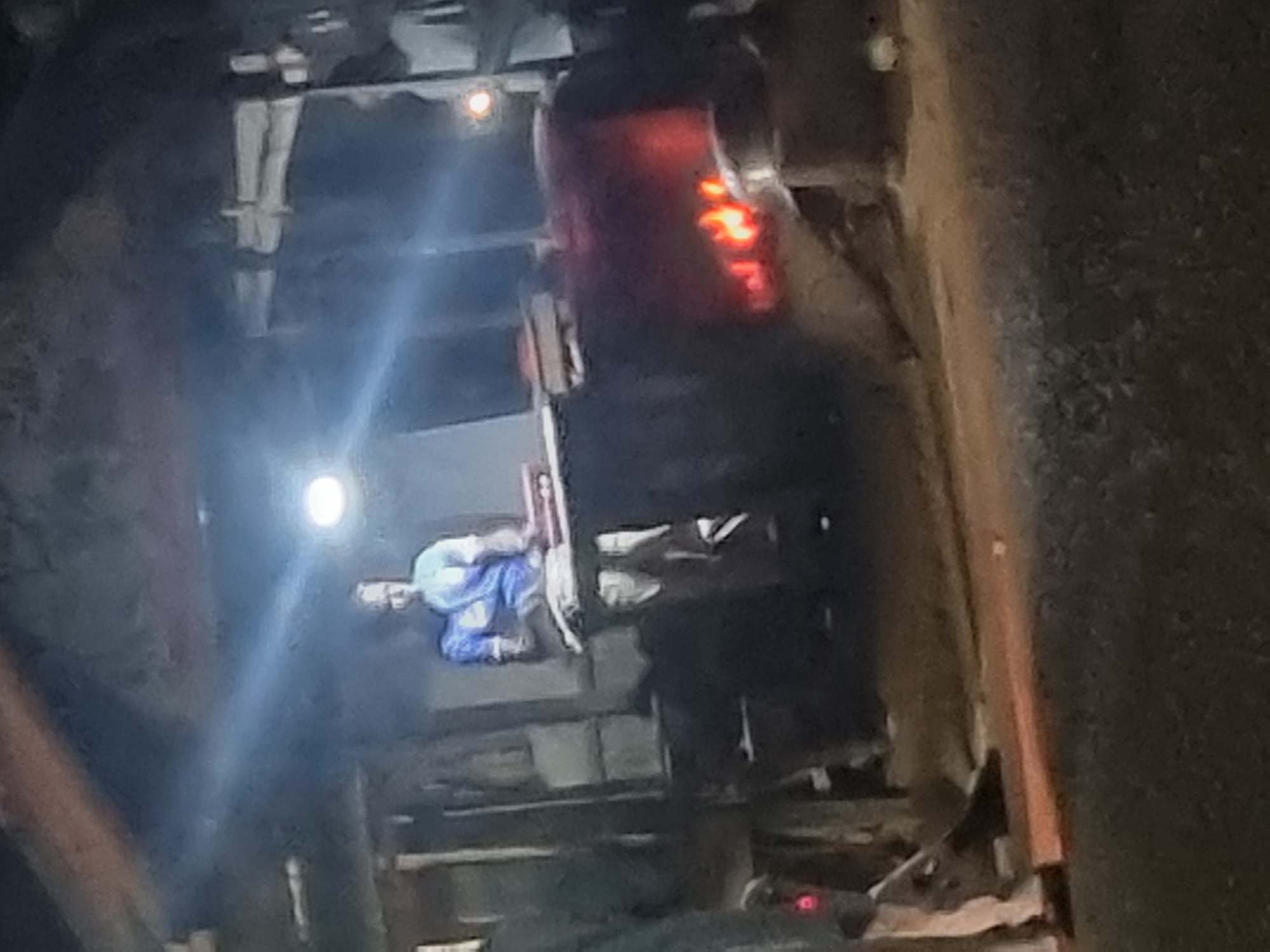 The image depicts an outdoor scene at night, possibly in Gwong, Nigeria, featuring a person wearing a blue shirt and white pants standing behind a wooden counter. The person appears to be engaged in some activity related to the items on the counter, which include what looks like books or papers. To the right of the person, a large, dark object is visible, with bright orange flames emanating from it, suggesting a fire or cooking area. The environment is dimly lit, with a bright light source illuminating the person and the counter from above. There are hints of structures in the background, possibly stalls or open-air buildings. The ground appears to be dirt or gravel. Another person is partially visible on the left side of the frame, wearing light-colored pants and a dark top, with a red light or object visible near their feet. The overall impression is that of a nighttime market or food stall setting.