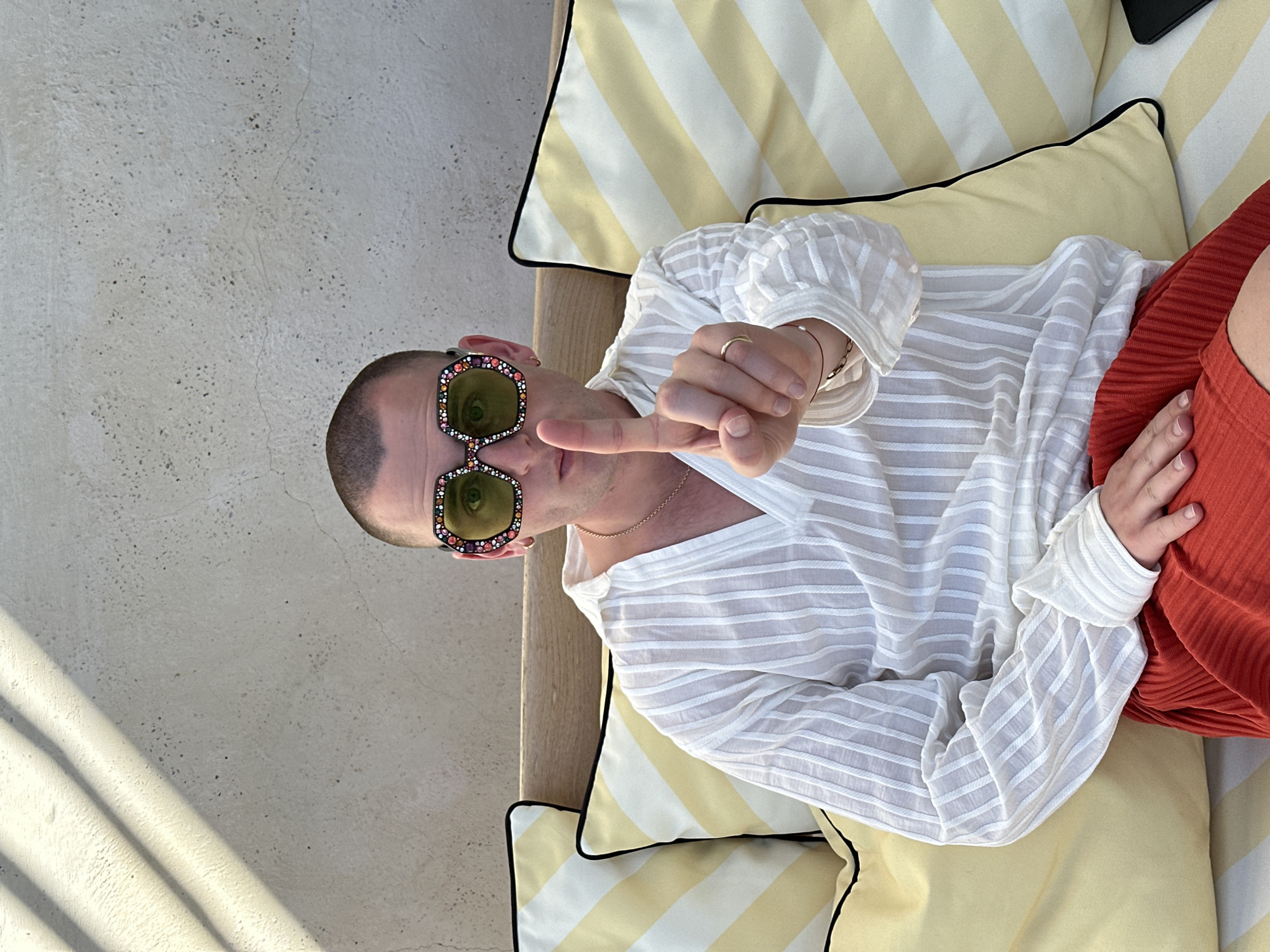 A person lies on a couch in Sant Josep de sa Talaia, Spain. The individual, who appears to be a man, has a shaved head and wears sunglasses with multicolored stones set in the frames. He wears a white, striped, sheer long-sleeved shirt. He is lying on yellow and white striped pillows with a black edge on a tan-colored couch. His right hand forms a pointing gesture. He wears a gold ring and bracelet. He is wearing red shorts. A stone-textured wall is in the background.