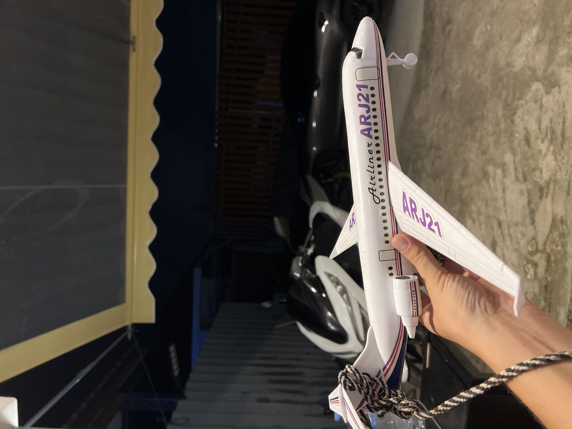 A person's hand holds a white toy airplane with purple lettering reading "Airliner ARJ21." The plane is a model of an ARJ21 aircraft. In the background, out of focus, is a motorcycle and the rear of a car. The scene appears to be outdoors, possibly in a garage or covered area, with a dark, corrugated metal fence and a retractable awning overhead. The ground is concrete.