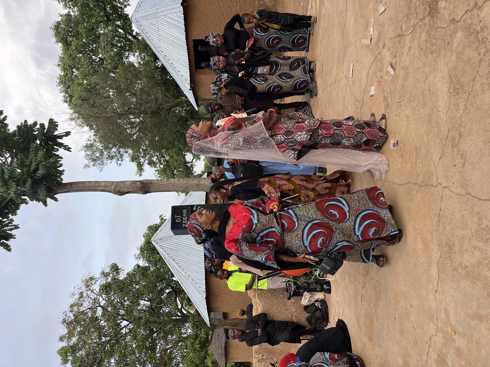 In the city of Jos, Nigeria, a group of people are gathered outdoors in what appears to be a rural or traditional settlement. The scene is characterized by several mud-brick houses with corrugated metal roofs. In the foreground, a woman in a vibrantly patterned, multi-colored dress and headscarf is the primary subject, looking to her right. To her left, another woman, also in a richly patterned dress and head covering, is partially visible. Behind them, several other individuals are standing and interacting. A prominent object in the mid-ground is a large black speaker with "DJ ZEENA" written on it, suggesting a gathering or celebration is underway. There is also some sound equipment visible. The ground is a light brown, earthy surface with some scattered currency notes. Trees surround the area, and the sky is overcast. The overall impression is of a community event or celebration taking place in a traditional Nigerian setting.