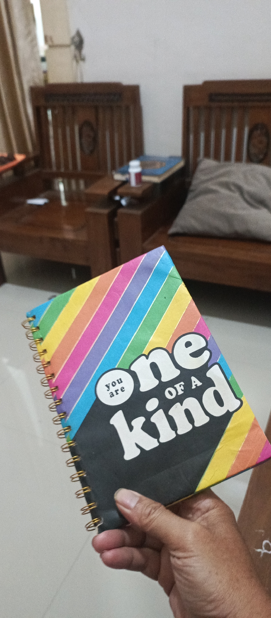 A hand holds a spiral-bound notebook with a colorful striped cover and the phrase "one of a kind" printed on the front. The notebook has a black background with white lettering, and the stripes are in rainbow colors. In the background, there is wooden furniture, including chairs and a table, suggesting an indoor setting. The scene is likely in Jatisari, Indonesia, based on the prompt.