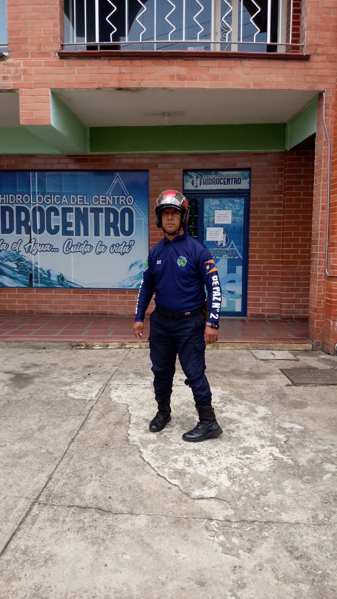 A man in a dark blue uniform, black boots, and a red and silver helmet stands outdoors in front of a brick building during the daytime in Tinaquillo, Venezuela. He is positioned centrally, facing forward with a neutral expression. His uniform includes a long-sleeved shirt with patches; one on his chest appears to be a circular logo, and another on his left sleeve reads "DE PAZ N° 2". He also has a device attached to his belt.
The building behind him is constructed of red brick. The ground floor features a large blue banner on the left, displaying "HIDROLÓGICA DEL CENTRO" and "HIDROCENTRO" prominently, with smaller text that reads "el Agua... Cuida la vida" (The water... Cares for life). To the right of the banner is a glass entrance door, also featuring the text "C.A. HIDROLOGICA DEL CENTRO" and "HIDROCENTRO". Above the ground floor, there is a second level of the brick building with windows protected by decorative white metal railings. The ground beneath the man's feet is a worn concrete surface with visible cracks and discolored patches. The overall lighting suggests clear weather conditions.