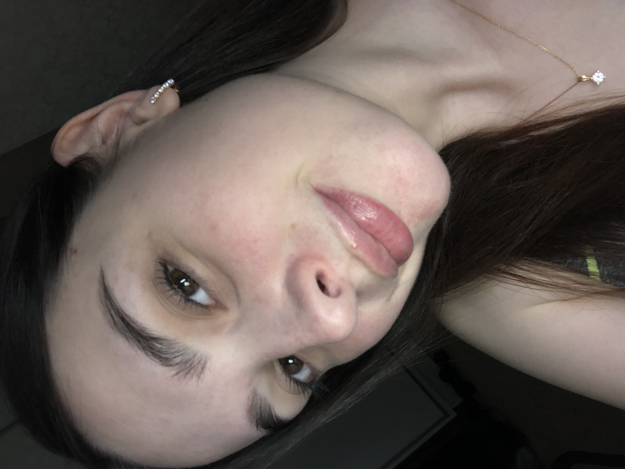 A close-up, angled selfie of a person with fair skin, dark brown hair, and brown eyes. The person is wearing an ear cuff with small stones on their left ear and a delicate gold necklace with a diamond pendant. The background is dark and out of focus, suggesting an indoor setting. There is no indication of a location in Noviy, Russia, or any city.