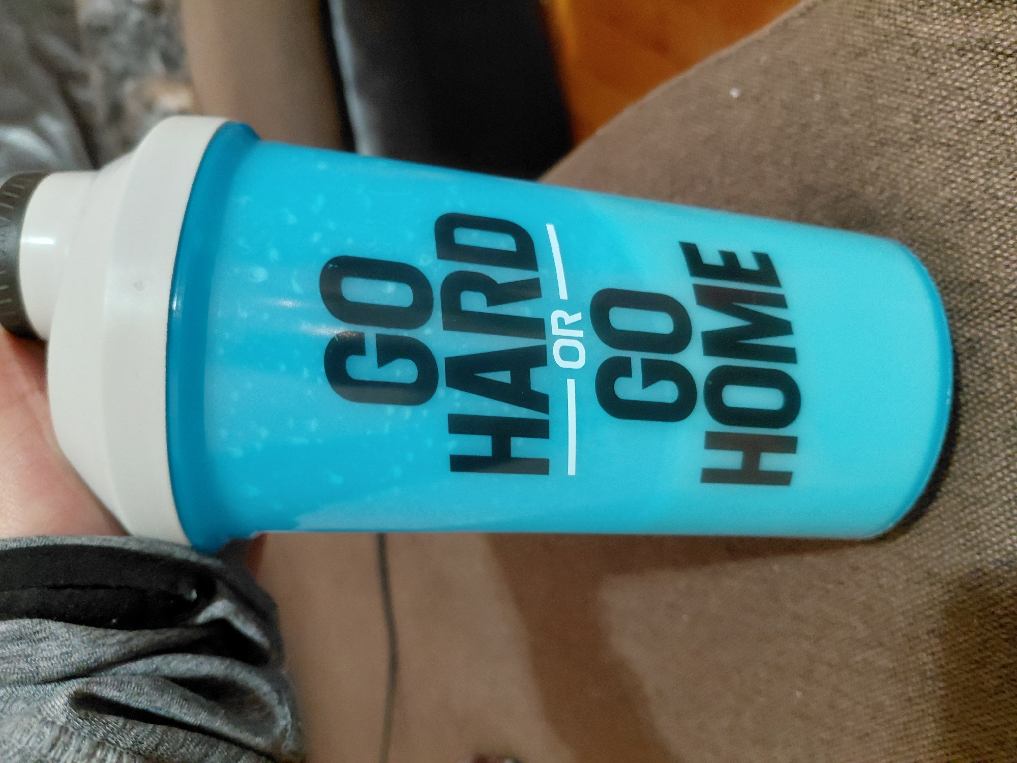 A close-up shot depicts a light blue shaker bottle with a white lid and black screw-on cap. The bottle has the phrase "GO HARD OR GO HOME" printed vertically in bold, black letters. The bottle is partially held by a hand with a gray sweater sleeve visible. The bottle rests on a brown upholstered surface. The location is unknown.