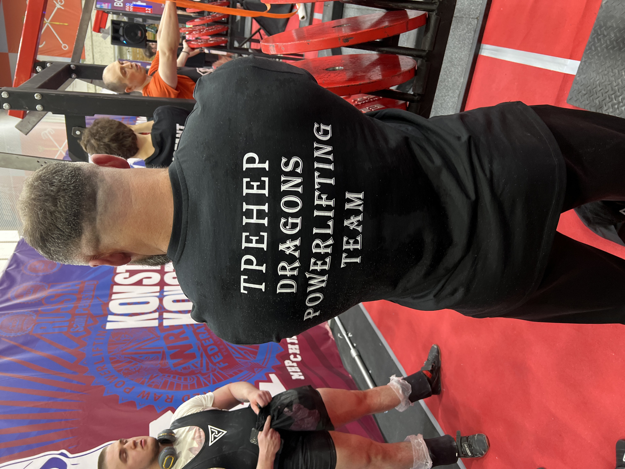 The image, oriented horizontally, shows a scene at a powerlifting event. A man in a black shirt with "ТРЕНЕР DRAGONS POWERLIFTING TEAM" printed on the back stands facing away from the camera. He has a closely cropped haircut with graying hair. To the left, another person is lying down, seemingly preparing for a lift, wearing a black singlet with white writing and symbols. An individual in an orange shirt is visible in the background, performing a pull-up on a rack with red weights nearby. A large backdrop is partially visible, featuring the words "KONST" and "Saint" above a blue and red emblem. The text "RAW POWERLIFTING FEDERA" is printed around the emblem. The event appears to be held in Saint Petersburg, Russia, based on the writing on the backdrop. The floor is covered in a bright red carpet.