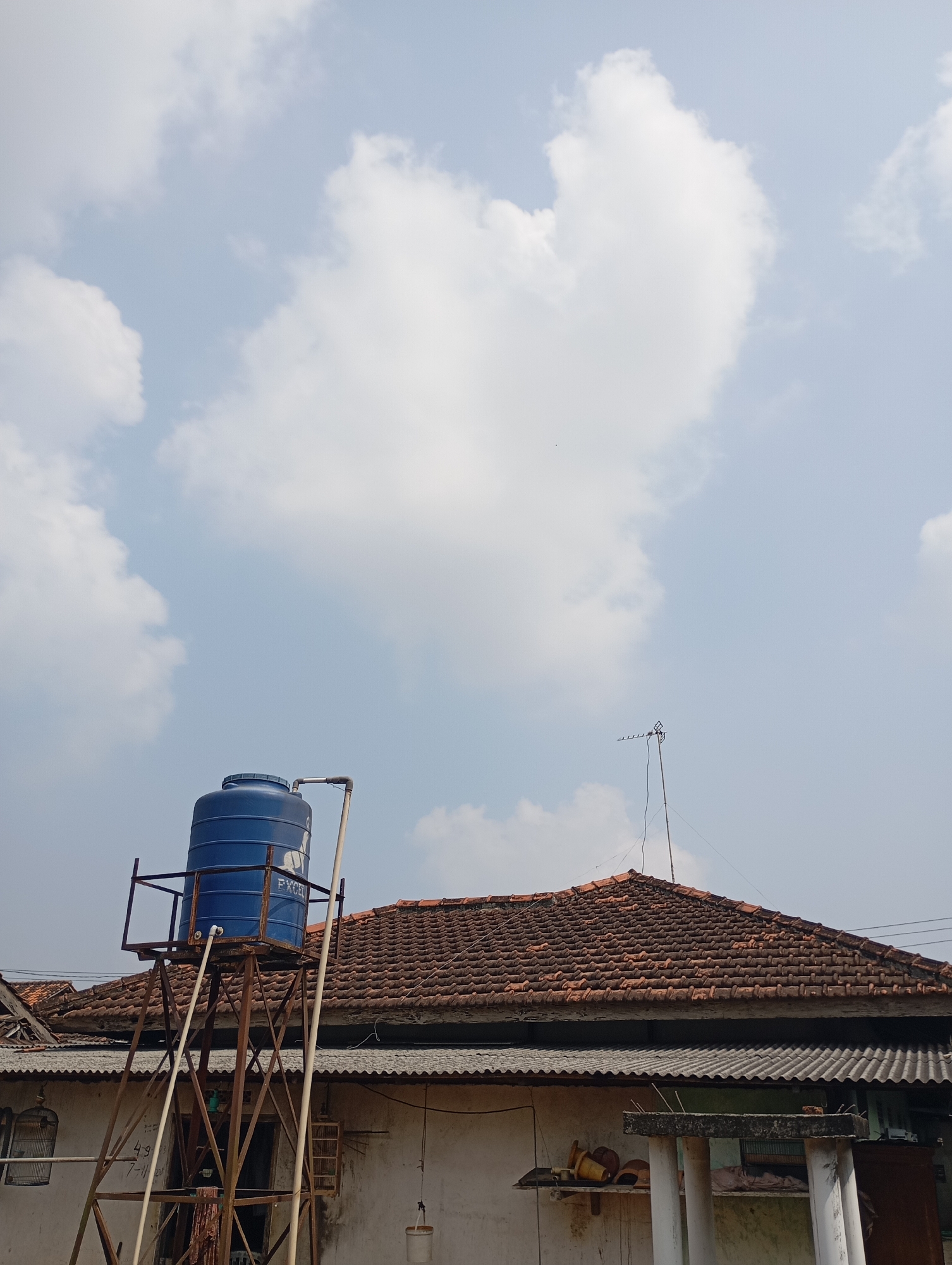 This is a vertical, eye-level outdoor photograph showing a blue water tank on a rusty metal stand in front of a residential building. The water tank is cylindrical with a rounded top and a cap. It is elevated by a brown metal framework. Pipes extend from the tank and down the side of the structure. Behind the water tank, a tiled roof with reddish-brown tiles covers a white building. A silver antenna stands on the peak of the roof. The sky above is a light blue with scattered white clouds. The scene is likely in a residential area of Metro, Indonesia, suggested by the style of the building and the presence of the water tank, a common utility in many Indonesian homes. There are no people visible in the image. The overall impression is one of a typical domestic setting with a focus on essential infrastructure.