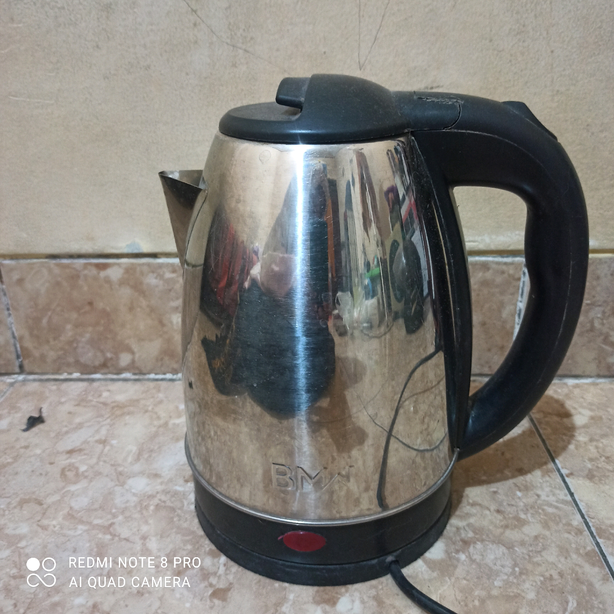 A stainless steel electric kettle with a black plastic handle and lid is placed on a tiled floor. The kettle has a visible "BMW" logo on its body and a red power button. The background consists of a beige tiled wall. The scene appears to be indoors, likely in a kitchen or living area. The image is overlaid with a "REDMI NOTE 8 PRO AI QUAD CAMERA" watermark in the bottom left corner, suggesting it was taken in East Jakarta, Indonesia. The lighting is natural, and the overall image quality is moderate.