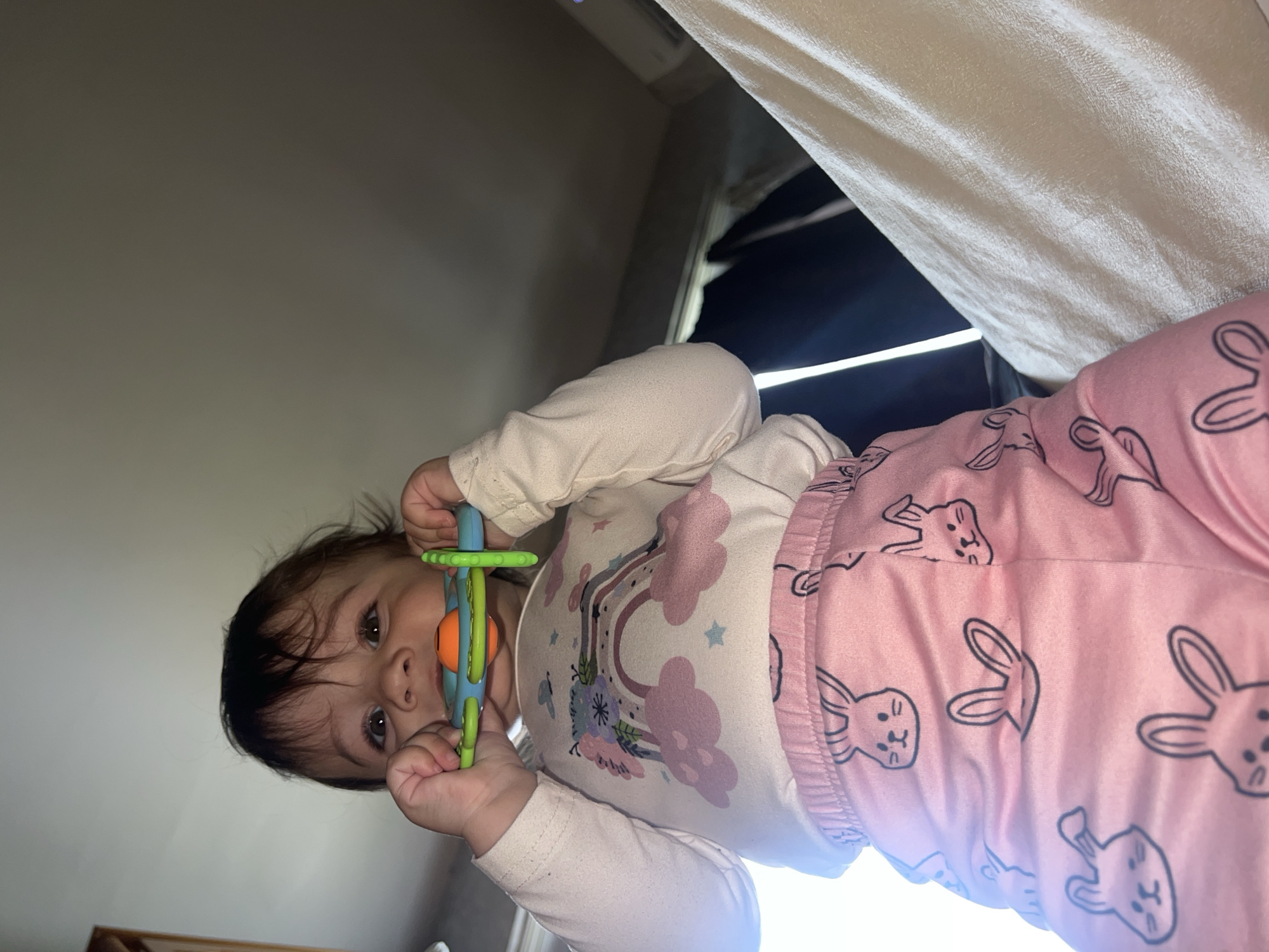 A baby is lying on its back, wearing pink pajamas with a bunny pattern and a long-sleeved white shirt with a rainbow design. The baby is holding a colorful toy in its hands. The background appears to be an interior room with a light-colored wall. The top right corner shows a white curtain and a glimpse of a dark area possibly outside a window. There is no indication of the city of Elena, Bulgaria.