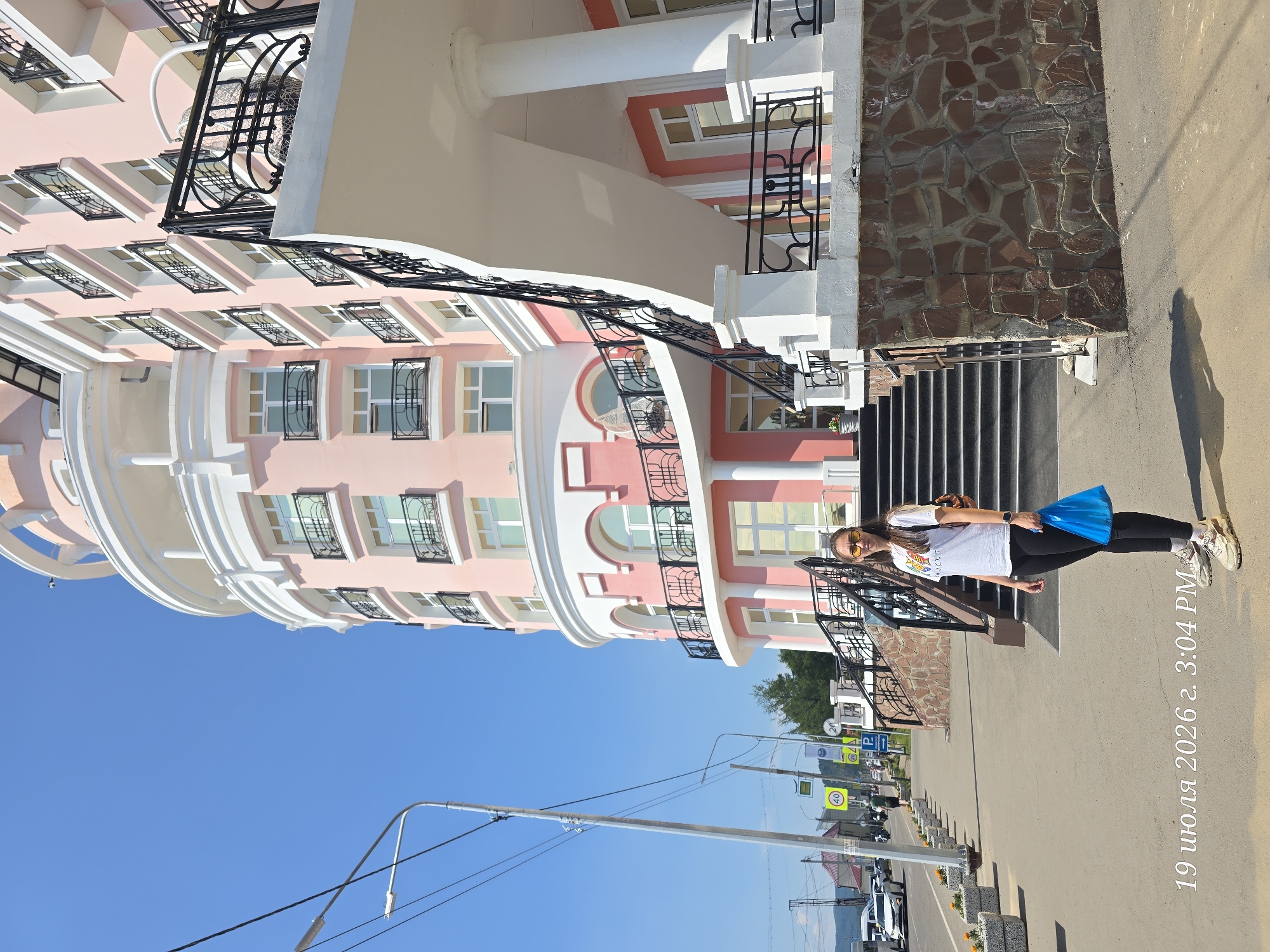 A young woman stands on a sidewalk in front of a pink building with white architectural details, possibly a hotel or apartment complex, in Listvyanka, Russia. She is wearing a white t-shirt, black leggings, sunglasses, and sneakers, and is carrying a blue plastic bag. The building has a curved facade, balconies with ornate black railings, and a prominent staircase leading up to an entrance. In the background, there is a clear blue sky, streetlights, traffic signs, cars, and other buildings. The timestamp "19 июля 2026 г. 3:04 PM" is overlaid on the bottom left of the image. The sidewalk has planters with orange flowers along the edge. The building's base is clad in stone.