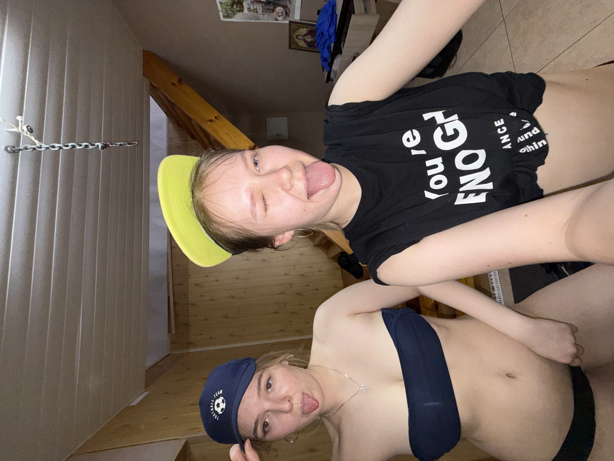 Two young women are posing for a selfie. The woman at the top of the image is wearing a black t-shirt with the words "You're ENOUGH and Change Nothing" and a neon yellow baseball cap. The woman at the bottom of the image is wearing a navy blue sports bra and a navy baseball cap with a soccer ball logo. Both women are sticking their tongues out and making playful faces. The background suggests they are indoors, possibly in an attic-like space with a sloped ceiling. There is a metal chain hanging from the ceiling and wooden support beams visible. The location is Zhigalki, Russia.