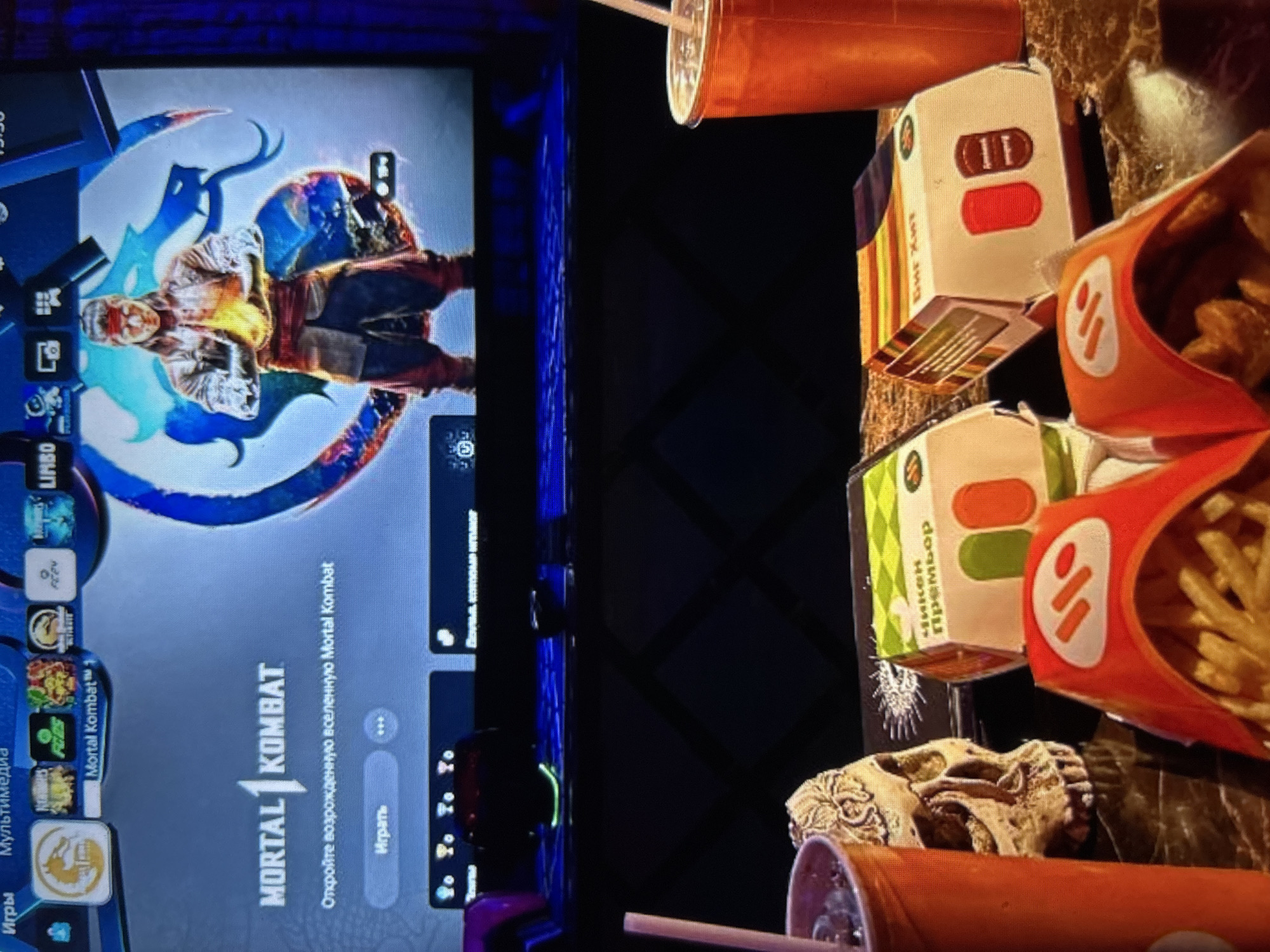 The image displays a screen showing the game Mortal Kombat 1, with the game's title and a description in Russian. Below the screen, a dark surface is visible, upon which several food items are placed: two fast-food boxes, one with "чикен премьер" (chicken premier) written on it, a french fry container filled with fries, a skull-shaped object, and a partially visible drink in an orange cup with a straw. Another orange cup with a straw is visible to the left. The background is dark and out of focus, suggesting an indoor, dimly lit environment. The overall scene appears to be a casual setting where someone is about to play a video game and consume fast food. The location is not identifiable from the image.