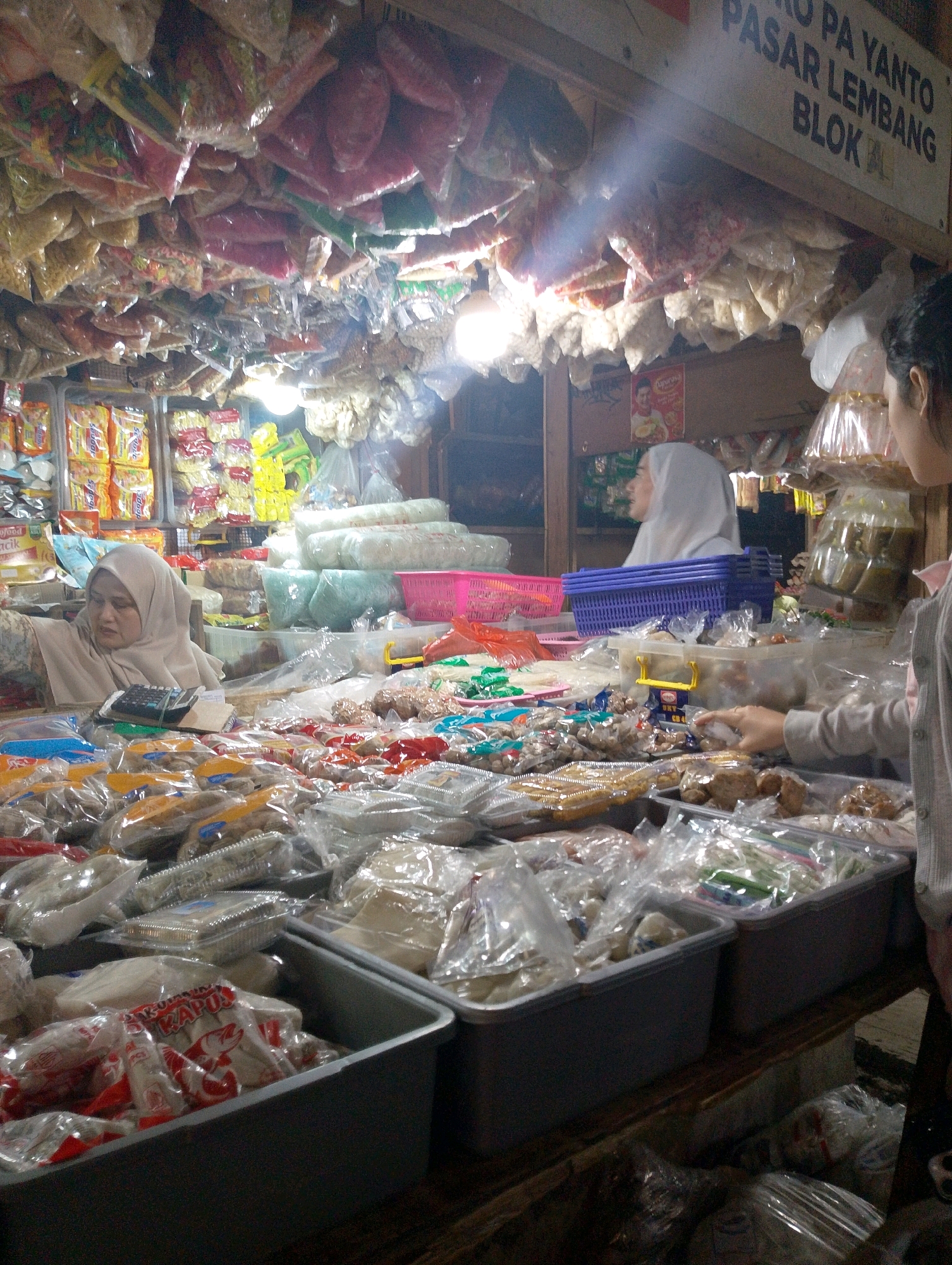 The image depicts a bustling market stall in Lembang, Indonesia. The stall is overflowing with various goods, primarily packaged food items, displayed in plastic bags and containers. Products are seen hanging from the ceiling and arranged on tables. Two women are present. One woman, wearing a hijab, is positioned behind a counter, interacting with the merchandise. Another woman, partially visible on the right, appears to be browsing or shopping. The market is dimly lit, with artificial lighting illuminating the products. A sign above the stall reads "PASAR YANTO LEMBANG BLOK". The scene suggests a typical Indonesian wet market environment, characterized by a wide assortment of goods and active commerce.
