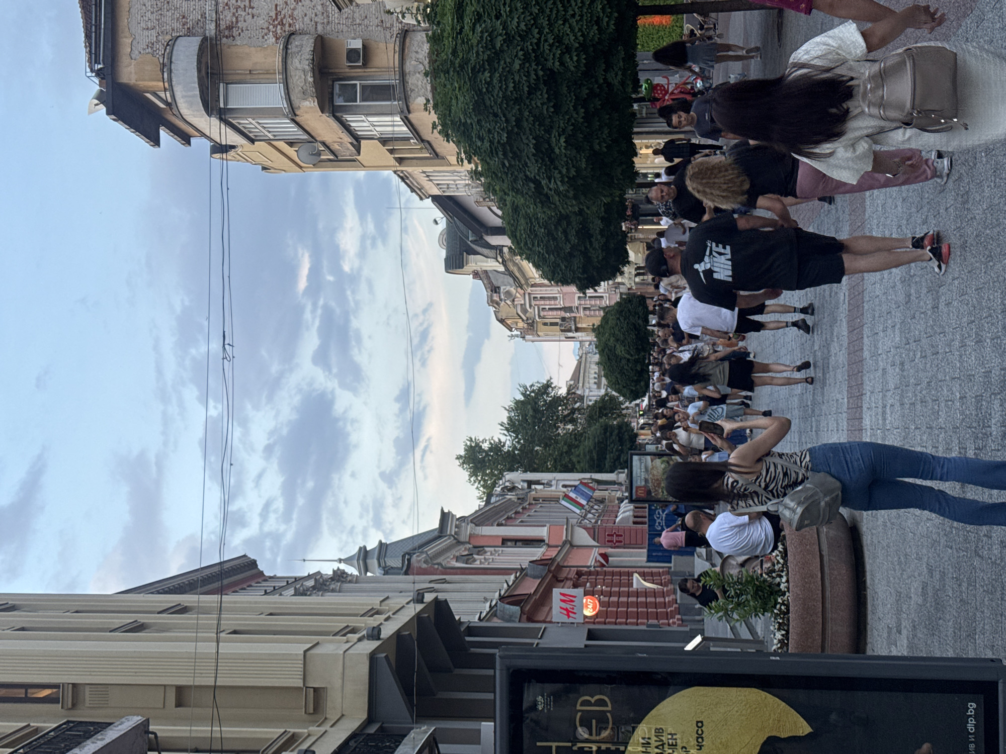 The image captures a bustling pedestrian street in the city of Plovdiv, Bulgaria, specifically Knyaz Alexander I Street, identifiable by the distinctive architecture and the visible H&M store. The scene is illuminated by soft, diffused light, suggesting late afternoon or early evening.
The street is paved with light-colored blocks and is lined on both sides by multi-story buildings (ranging from three to five floors) exhibiting diverse architectural styles, including classical elements, ornate balconies, decorative cornices, and pitched roofs. Building facades are painted in various pastel shades of beige, pink, yellow, and white. Utility wires are visible stretched across the street against a cloudy sky.
On the left side, a large, dark vertical advertising stand with Cyrillic text is partially visible in the foreground, displaying "dtp.bg" at the bottom. Further down the street on the left, an H&M store is visible with a prominent red "H&M" sign and a smaller "24/7" sign below it. Three national flags—Hungarian, Bulgarian, and another with a green stripe (possibly Italian or Mexican, difficult to discern precisely)—are displayed from a building facade on the left. On the right, a building features a rounded balcony, a satellite dish, and an air conditioning unit. Several neatly trimmed, spherical trees line the center of the pedestrian area, interspersed with large, round planters containing small bushes or plants. A McDonald's sign is partially visible further down the street on the right.
Numerous people are present, walking in various directions along the street, indicating a busy public space. Most individuals are adults and young adults dressed in casual attire such as t-shirts, jeans, shorts, and dresses. In the immediate foreground, a woman with her back to the viewer wears a zebra-print tank top, blue flared jeans, and a grey backpack, holding a smartphone up to take a photograph. Behind her and slightly to the right, a man in a black t-shirt with a white "NIKE" logo and black shorts walks hand-in-hand with a woman in pink pants. Further to the right and slightly closer, another woman in a white dress and a grey backpack holds hands with an unseen individual to her right. People are observed walking, conversing, and sitting on benches near the planters.