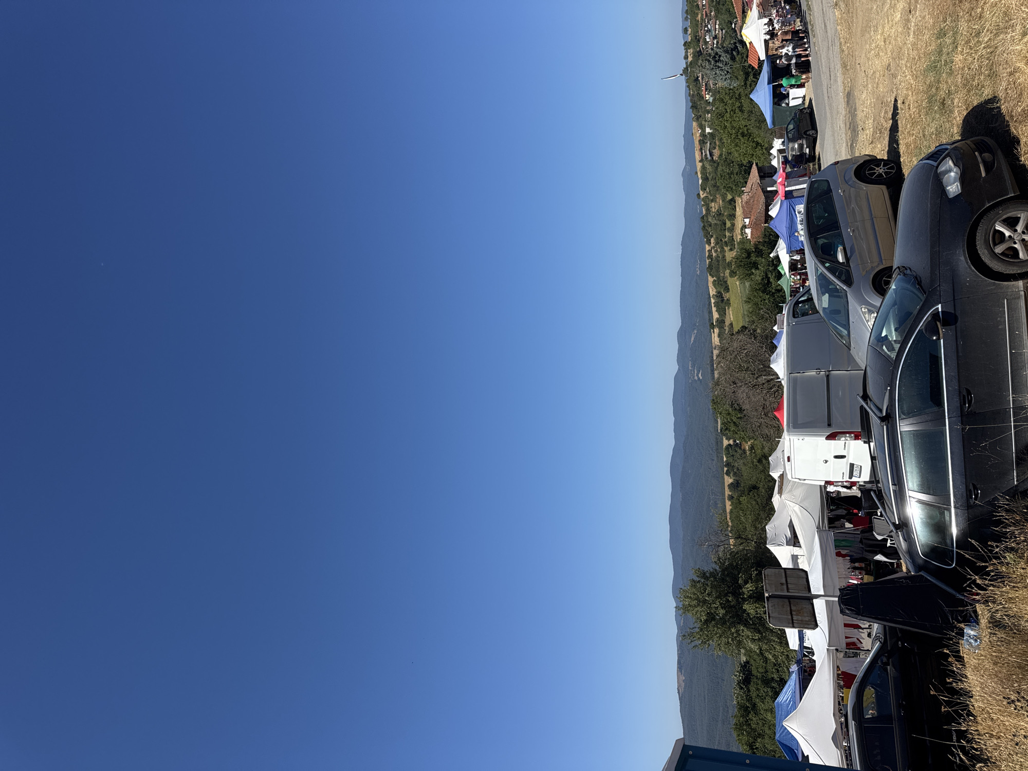 The image depicts an outdoor market event in the village of Zheravna, Bulgaria. The scene is characterized by numerous stalls set up under various colored tents, predominantly white and blue. These stalls appear to be selling merchandise, with items visible on display. Several vehicles are parked around the market area, including black cars, a silver minivan, and a white van. In the foreground, tall, dry grass is present. In the background, rolling hills covered with trees and vegetation form the landscape, with houses and buildings visible amongst them, suggesting a village setting. The sky is a clear, bright blue. There are people browsing the stalls and interacting at the market. The overall atmosphere suggests a sunny day and a community event.