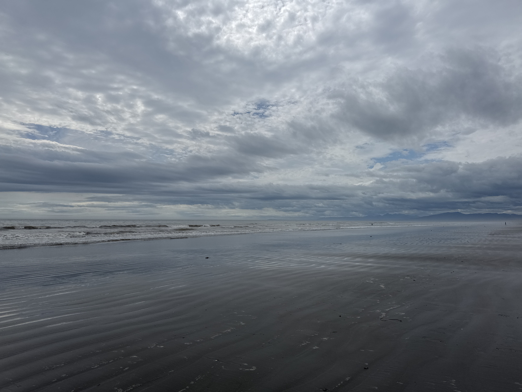The image depicts a wide, wet, sandy beach with ripples on the surface, likely caused by receding tide. The sea is calm with gentle waves breaking on the shore. The sky is overcast with dramatic clouds. In the distance, a low-lying, hazy mountain range can be seen. There are a few indistinct human figures present on the beach. This scene is consistent with the coastline near Poronaysk, Sakhalin Oblast, Russia, known for its expansive beaches and often cloudy skies.