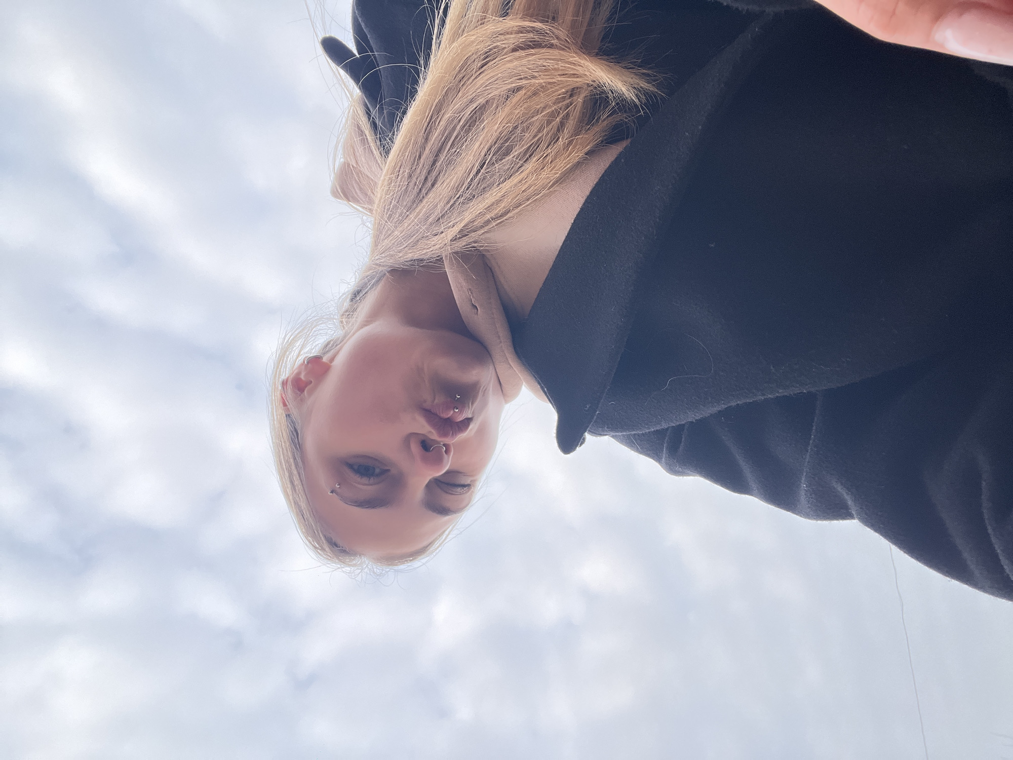 A low-angle, head-and-shoulders image of a light-skinned individual with blonde hair, taken outdoors in Фоки, Russia, against a cloudy sky. The subject is looking down towards the camera with a puckered lip expression. Visible piercings include a silver barbell on the left eyebrow, a silver ring on the nose, and two silver labret piercings on the upper lip. The individual is wearing a dark, possibly black, textured outer garment over a light beige or light brown inner layer. A portion of the individual's right hand or fingers is visible in the lower right corner.