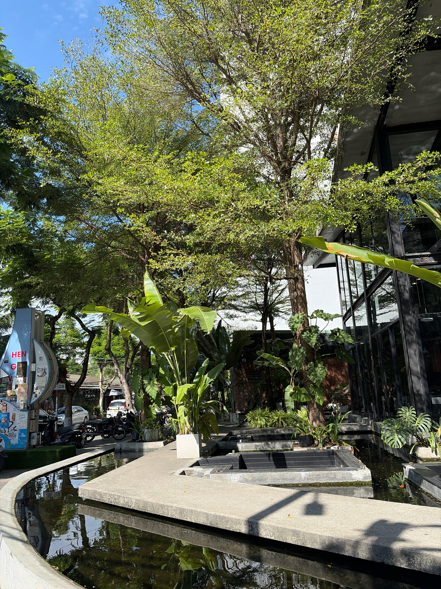 The image captures an outdoor scene in Da Nang, Vietnam, likely a cafe or a public space with a modern architectural design. In the foreground, a curved concrete pathway borders a reflective pool of dark water. The pool's surface mirrors the surrounding lush greenery and the clear blue sky. Beyond the pool, a paved walkway leads towards a building with large glass windows and black metal framing, suggesting a contemporary aesthetic. Several trees with dense green foliage are prominent, casting dappled shadows on the ground. Large-leafed tropical plants, such as banana plants, are strategically placed, adding to the verdant atmosphere. To the left, a blue and white cylindrical informational kiosk is visible, adorned with various posters and stickers, indicating it is a public area. Several parked motorcycles and a car are present in the background, hinting at an urban setting. The lighting suggests it is daytime, with bright sunlight illuminating the scene.
