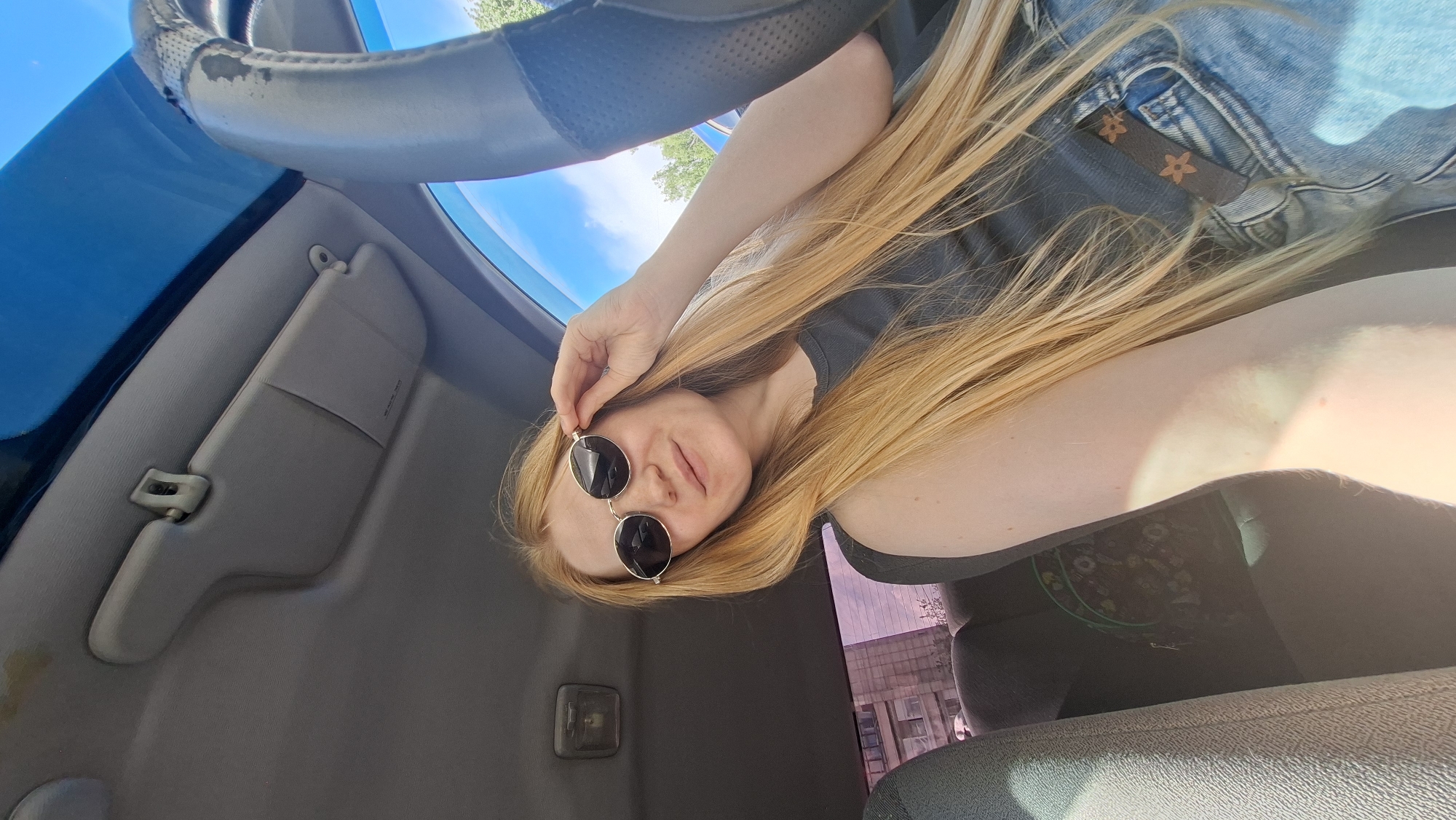 The media file shows a young woman with long, blonde hair and sunglasses, sitting in a car. She is wearing a grey tank top and denim shorts. The sun is shining, casting bright light into the car. The image appears to be taken from a low angle, looking up towards the woman and the car's interior. The car's sun visor is visible, as is the steering wheel. The background shows a glimpse of trees and sky through the car's windows, suggesting it is daytime and outdoors. The woman has a relaxed expression, and the overall mood is casual and sunny. There are no visible text elements in the image. The location context is Chaikovsky, Russia.