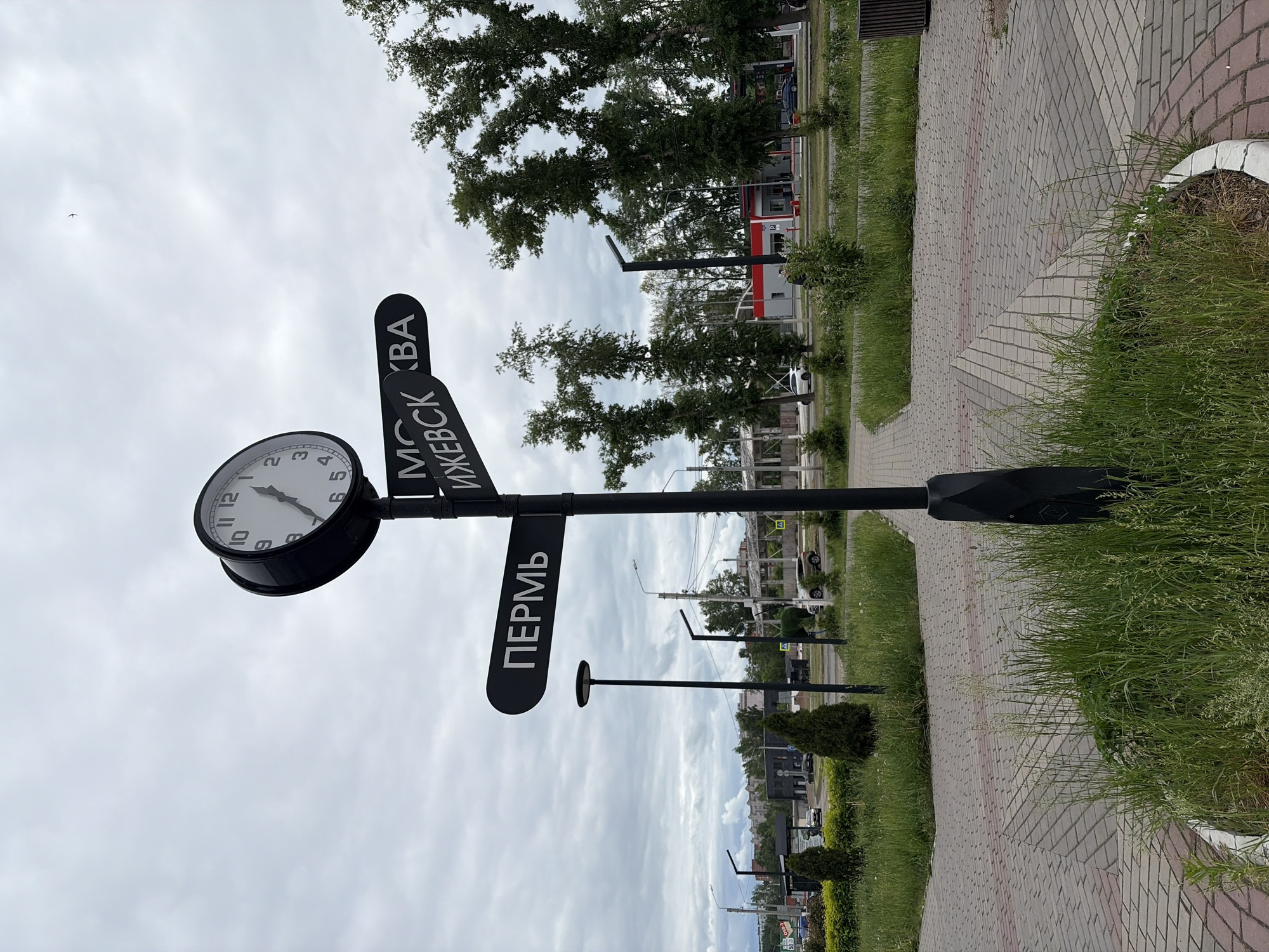 The image shows a clock and signpost in the city of Chaikovsky, Russia. The signpost has a clock at the top, followed by signs pointing in different directions labeled with city names: "Moscow," "Izhevsk," and "Perm." The signpost is black and stands on a brick-paved walkway. A planter with green grass surrounds the base of the post. Behind the walkway are patches of grass, shrubs, trees, and buildings. Streetlights are visible in the background. The sky is overcast.