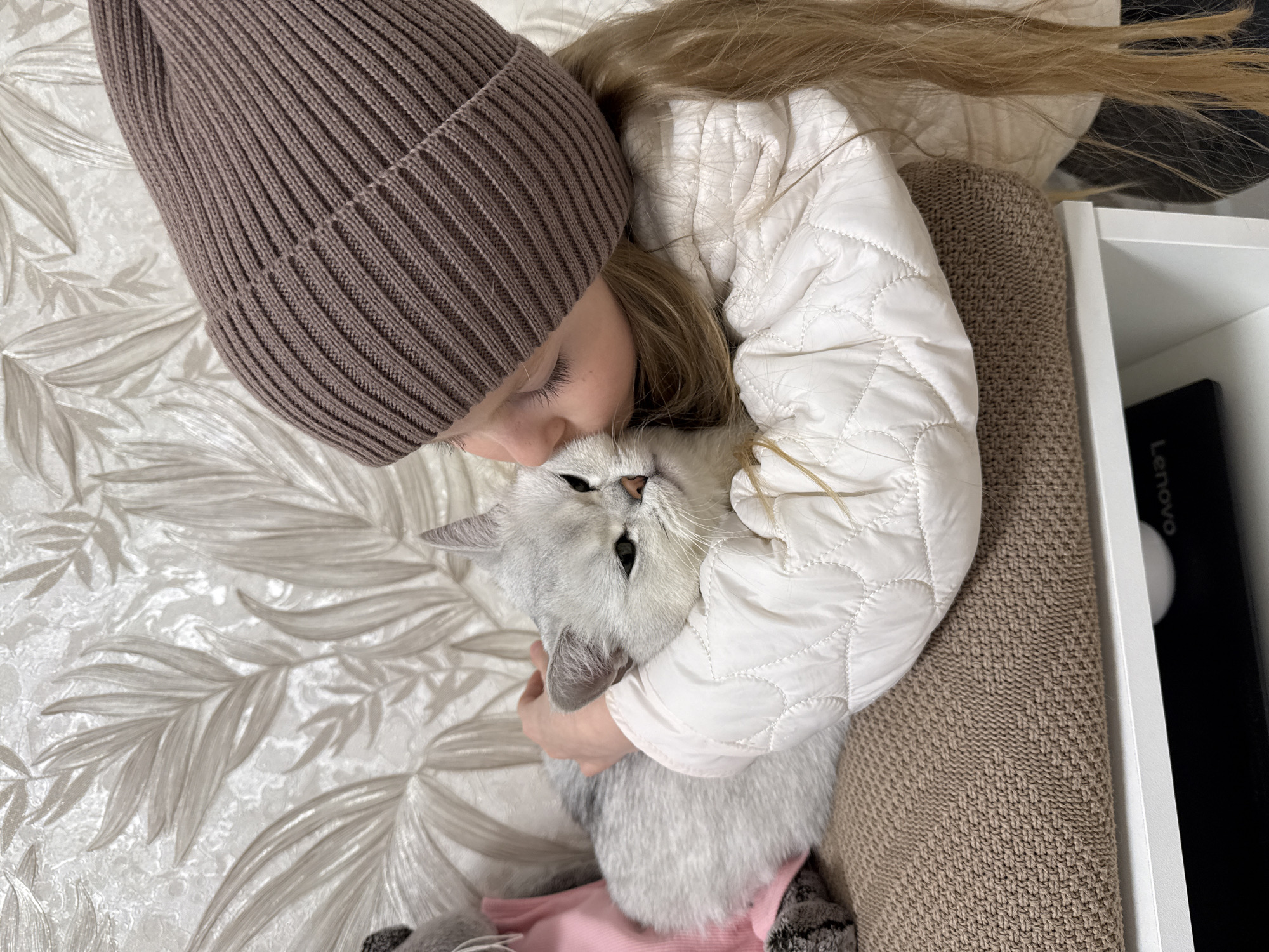 A young girl with long blonde hair wearing a brown knit hat and a white quilted jacket is cuddling a silver tabby cat with green eyes. The girl is lying on a bed with a leafy patterned bedspread. She has her arms wrapped around the cat, and her face is close to the cat's head, as if she is kissing it. The cat looks directly at the camera. The scene is indoors, and there is a brown textured couch or chair to the right, partially obscured. A white shelf with a black Lenovo laptop or notebook is visible in the upper right corner. The image does not provide enough information to determine if this is Izhevsk, Russia.