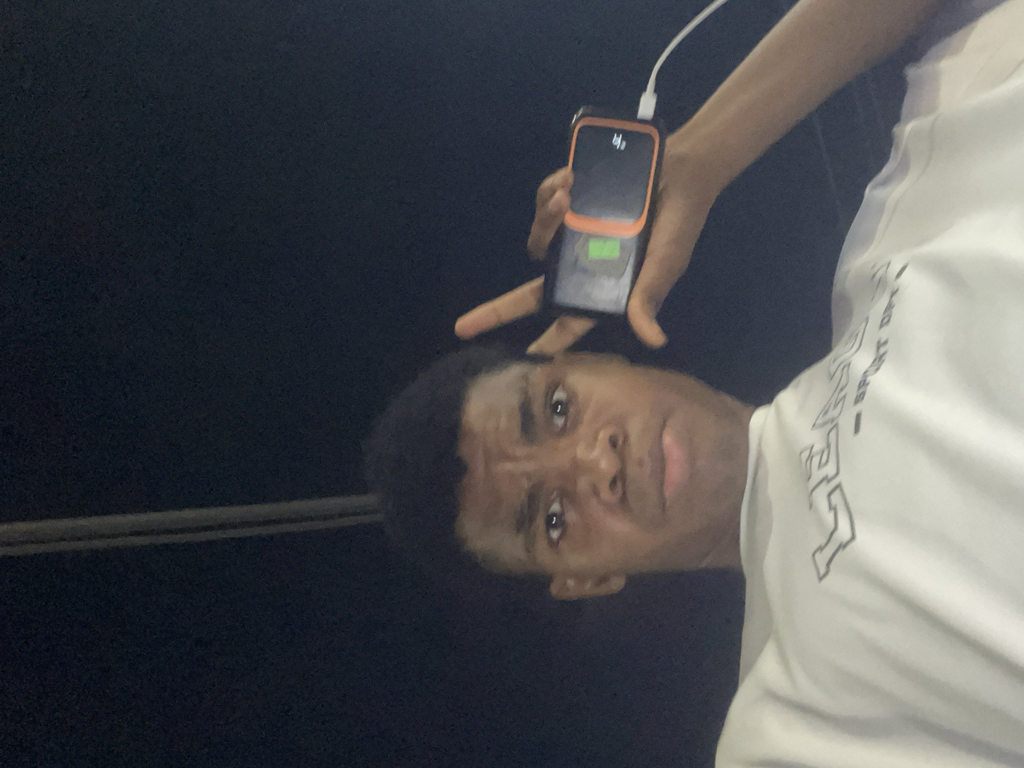 A person is holding a charging mobile phone. The background is a dark, indeterminate area, possibly outside at night. The person's face and torso are visible; they are wearing a white shirt. A cable connects the phone to a power source. There's a pole in the background. The location of Ado, Nigeria, is not evident in the image.