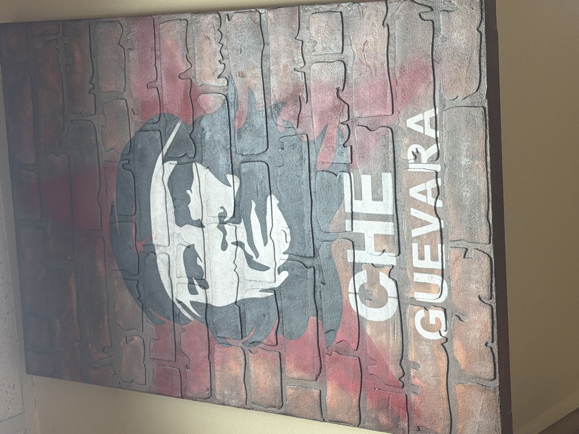 The image shows a large, framed piece of art, possibly a mural or a stretched canvas, depicting a stylized portrait of Che Guevara. The artwork is set against a background that mimics a brick wall, with red and grey bricks. Che Guevara's face is rendered in black, white, and grey tones. Below the portrait, the words "CHE GUEVARA" are printed in large white sans-serif font, also designed to appear as if painted on the brick surface. The artwork is mounted on a wall, with the top edge of the frame visible against a textured ceiling. The bottom portion of the image shows a beige-colored wall below the artwork. There are no people visible in the image. The context of Maloyaroslavets, Russia, is not visually supported by the image content.