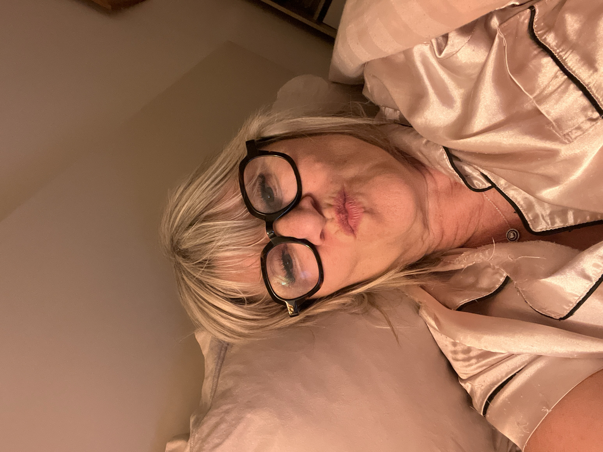 A medium-shot, eye-level selfie captures an adult female from the chest up, lying in a bed within an indoor setting in Chambœuf, France. The woman, appearing to be middle-aged, has shoulder-length blonde hair with bangs and wears dark-rimmed glasses, which reflect ambient light. Her lips are puckered in a distinct kissing gesture, with slight creasing visible around the mouth. She is dressed in a light pink or champagne-colored satin pajama top with dark piping along the collar and appears to wear a silver necklace with a circular pendant containing a central stone. Behind her head to the left is a light pink pillow, and striped bedding in a similar hue is visible on her right side, covering her torso. The background consists of a plain, light peach-toned wall or ceiling, illuminated by warm, dim artificial light. No other individuals or specific room features are identifiable.
