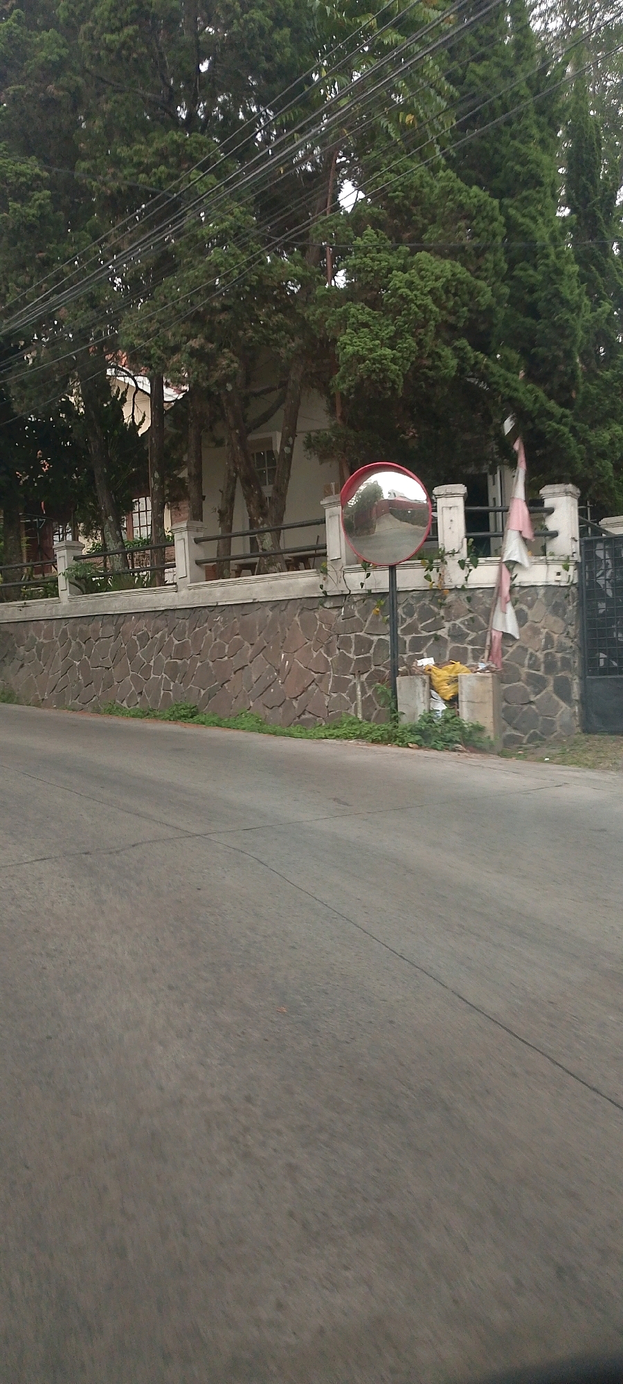 The image captures a street scene in Wangunsari, Indonesia, featuring a road in the foreground and a stone wall with a white fence and lush trees in the background. A convex mirror, commonly used for traffic safety, is positioned on the right side of the road, mounted on a black pole. Behind the wall, a white building with windows is visible, partially obscured by the trees. Several power lines run horizontally across the upper portion of the image, weaving through the dense foliage of the tall trees. A trash bin is situated near the mirror, with some yellow and white bags of refuse placed beside it. A dark metal gate is partially visible on the far right. The overall atmosphere suggests a residential or semi-residential area with a slightly overcast sky.
