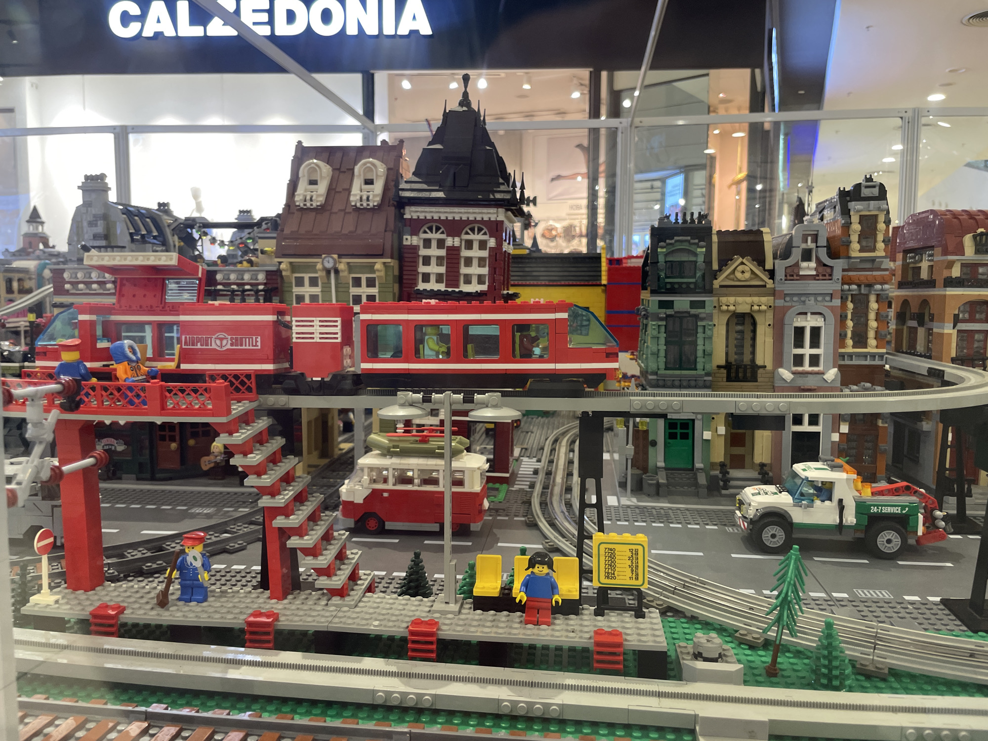 The image displays a detailed Lego city model, likely a store window display, given the prominent "CALEDONIA" signage visible at the top. The model depicts a train station with a red train, a red and white bus, a tow truck, and several Lego figures. The buildings are varied, featuring multiple stories, intricate facades, and pitched roofs, suggesting an urban environment. Train tracks weave through the scene, connecting various elements of the city. Yellow Lego benches are present at the train platform. The scene is well-lit, with reflections from overhead lights visible on the glass enclosure of the display. The setting does not provide any specific details to infer the location as Sofia, Bulgaria.