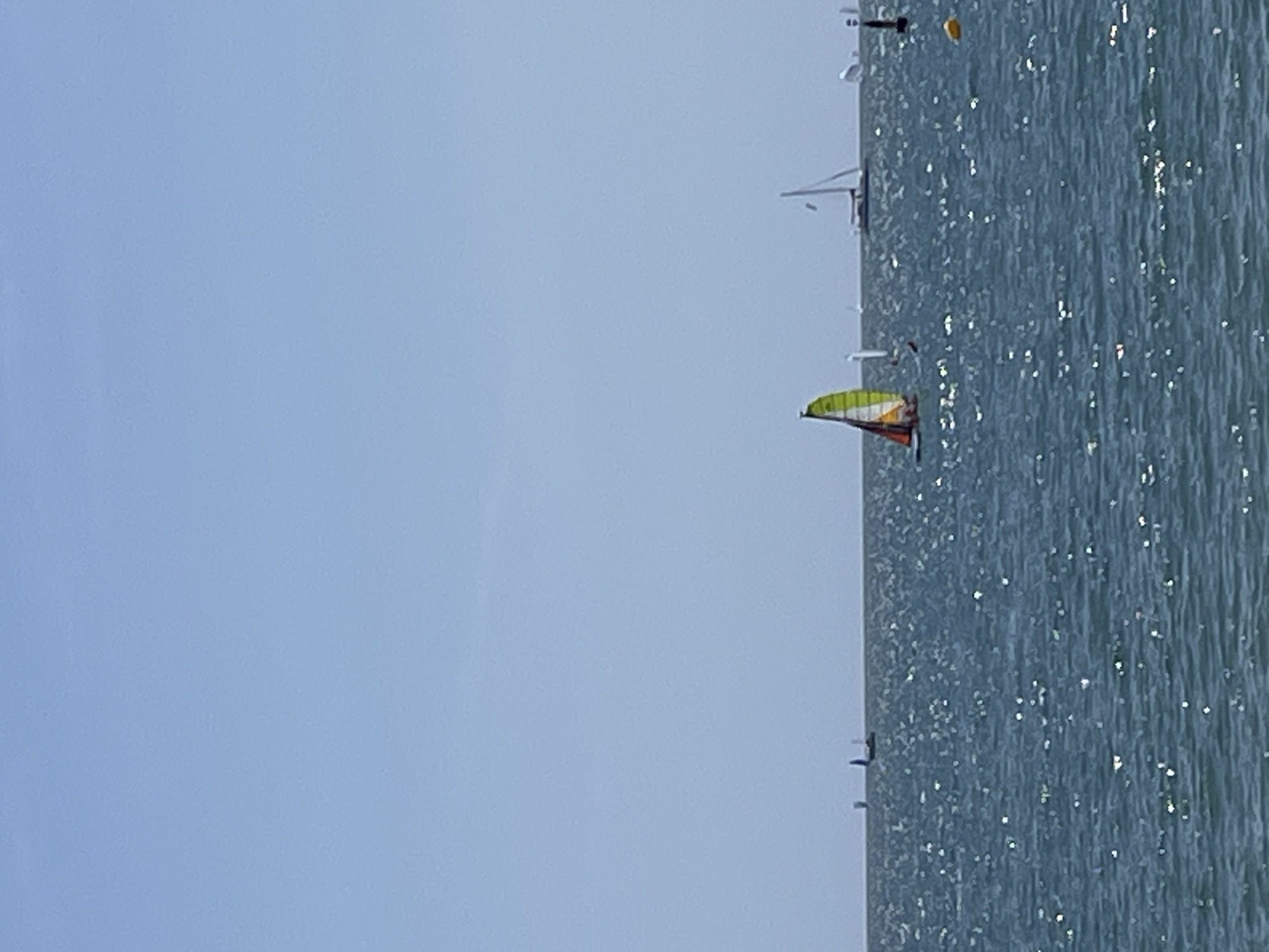 The image depicts a body of water, possibly the sea, under a clear, light blue sky. The water surface is slightly rippled, with light reflecting off the waves, creating a shimmering effect. Several boats are visible on the water, including one with a colorful sail that is predominantly yellow and green. Other boats appear to be sailboats. Given the presence of boats on the water under a clear sky, the scene likely takes place during the day. While without additional information, the location cannot be specifically confirmed, the presence of sailboats could suggest that the image was taken along the coast of Barcelona, Spain.