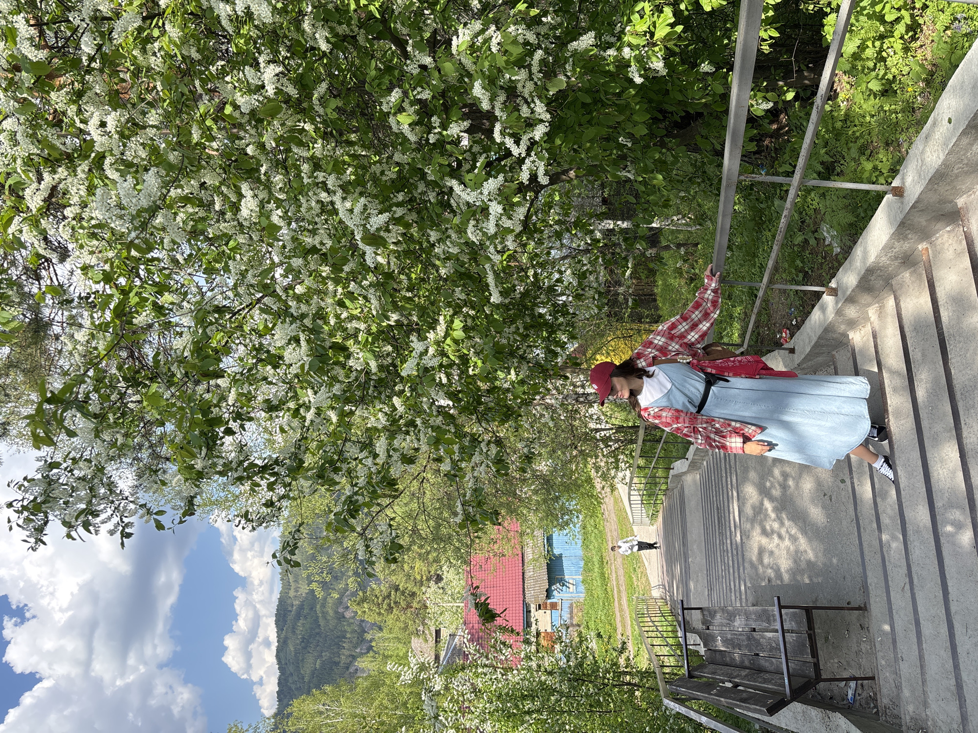 A full-length shot shows a woman descending a concrete staircase in Овсянка, Russia. She wears a blue dress with a black belt, a red and white plaid shirt draped over her shoulders, a red baseball cap, and black and white sneakers. She holds onto a gray metal railing as she walks down the stairs. The background features a blossoming tree with white flowers, a partly cloudy sky, and a distant view of buildings and a mountain. A bench is visible near the bottom of the staircase, and another person can be seen in the distance. The setting appears to be a public space within the city.