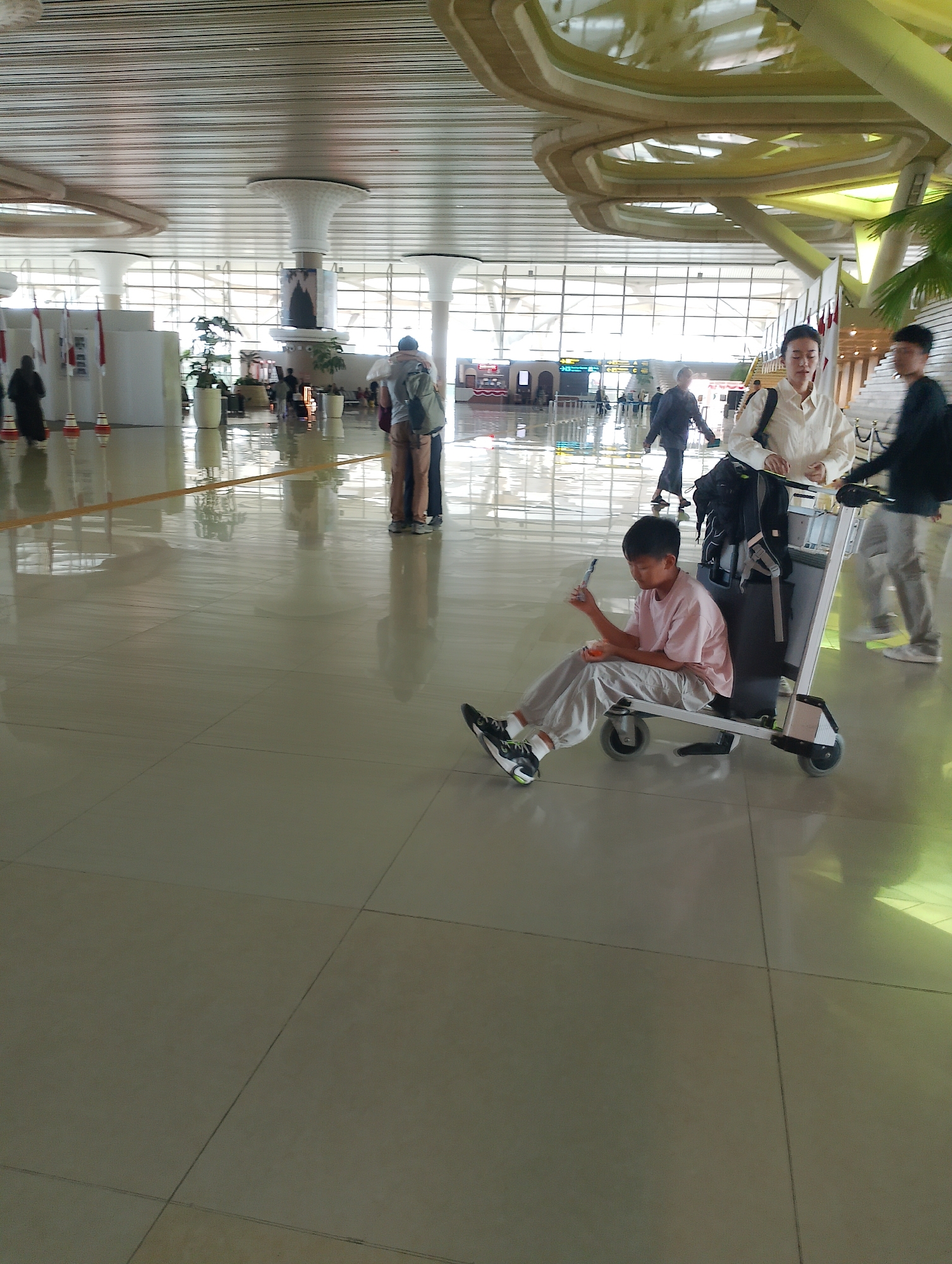 An outdoor shot captures the interior of an airport terminal, likely in Palihan, Indonesia, with a focus on a young boy seated on a luggage cart. The boy, wearing a light pink t-shirt and light-colored pants, is holding a small object in his right hand. He is positioned in front of the cart, his legs bent, and his body angled slightly to his left. Beside and behind him, a woman in a white top stands, with a backpack on the cart. Another person in a dark jacket is visible to the right, walking away from the camera. The airport interior features a high, modern ceiling with intricate lighting fixtures and a wall of windows in the background. The polished floor reflects the overhead lights and architectural elements. Several flags and potted plants are visible in the distance, along with other travelers moving through the terminal.