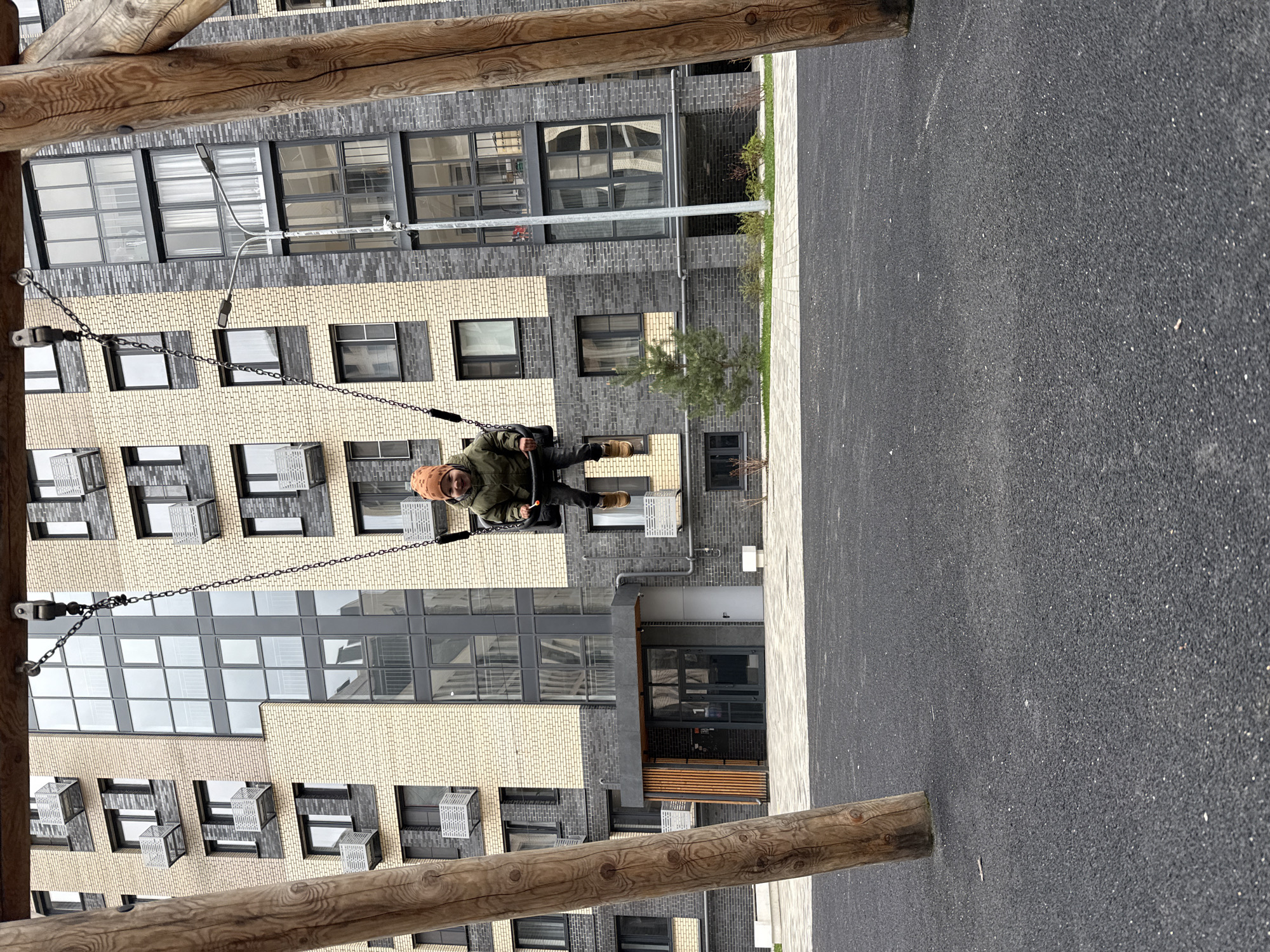 A person in a green jacket and brown hat is sitting on a swing in a playground in Rusanovo, Russia. The playground has a black rubber surface. Behind the swing is a multi-story apartment building with brick facade in alternating colors of tan and gray. Windows are visible on all floors. The swing is constructed of wooden beams and metal chains. The scene is well-lit, indicating daytime.